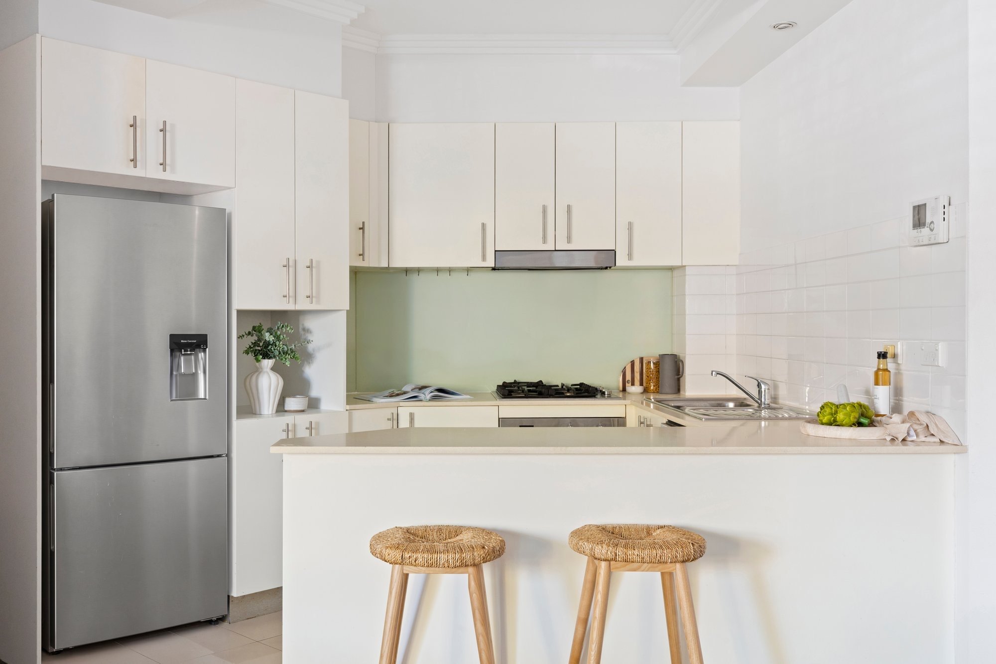 Modern white kitchen with stainless steel refrigerator, white cabinets, a small countertop with a potted plant, a bowl, a book, and some food items, including grapes and a bottle of oil, on the kitchen island with two wooden stools.
