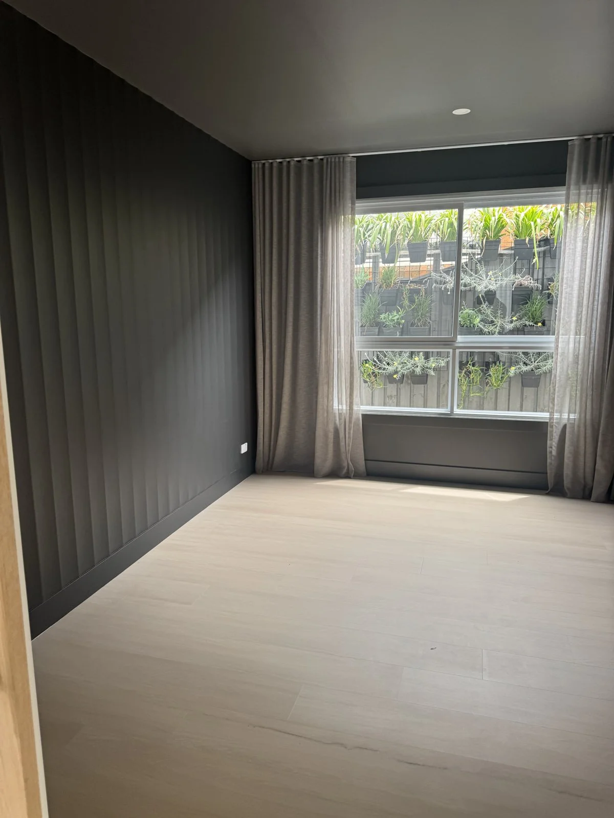 Empty room with black accent wall, beige curtains, and a window with potted plants outside.