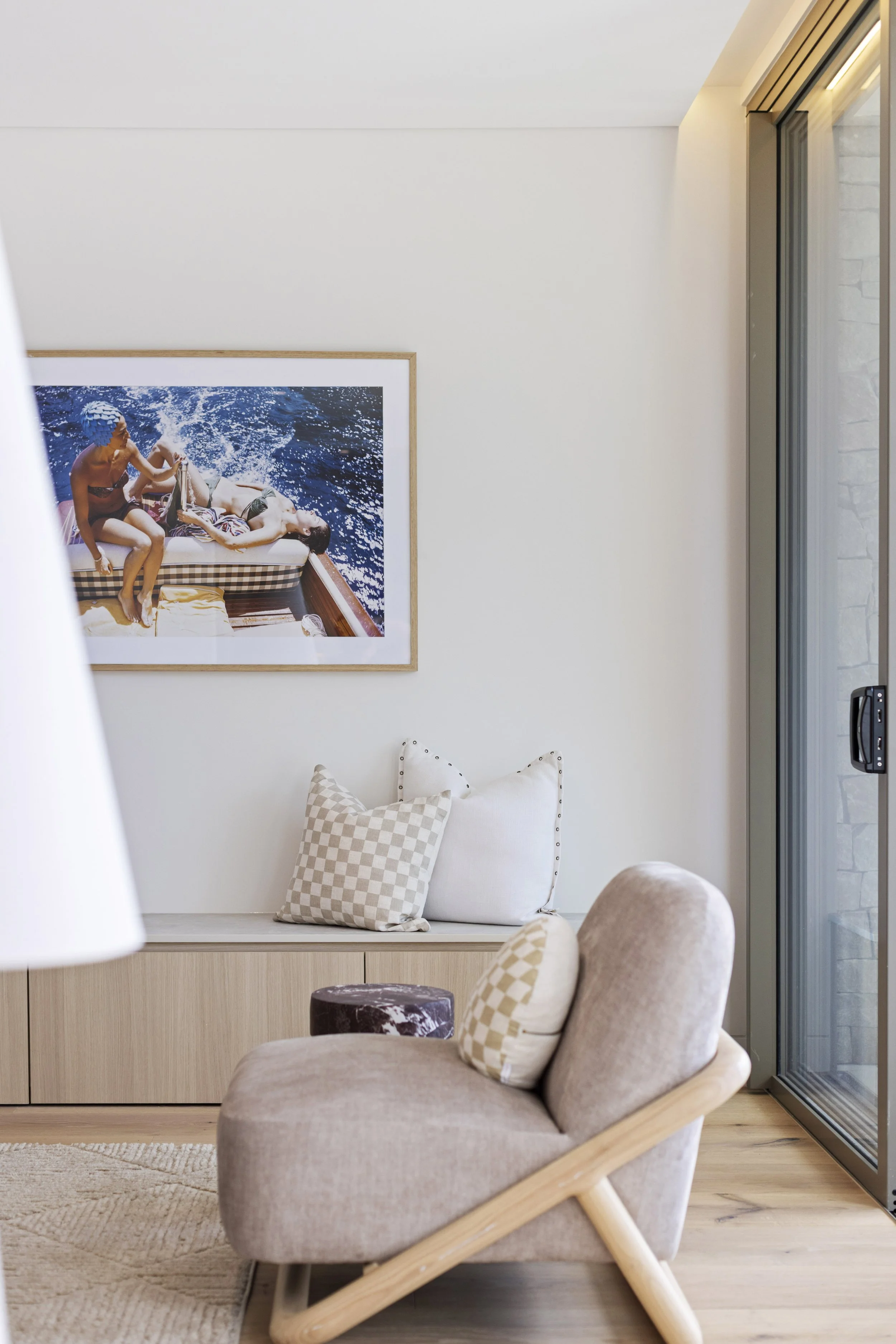 Modern living room featuring a beige armchair with beige and checkered pillows, a built-in wooden cabinet, a large framed photograph of people on a boat, and large glass sliding doors to the right.
