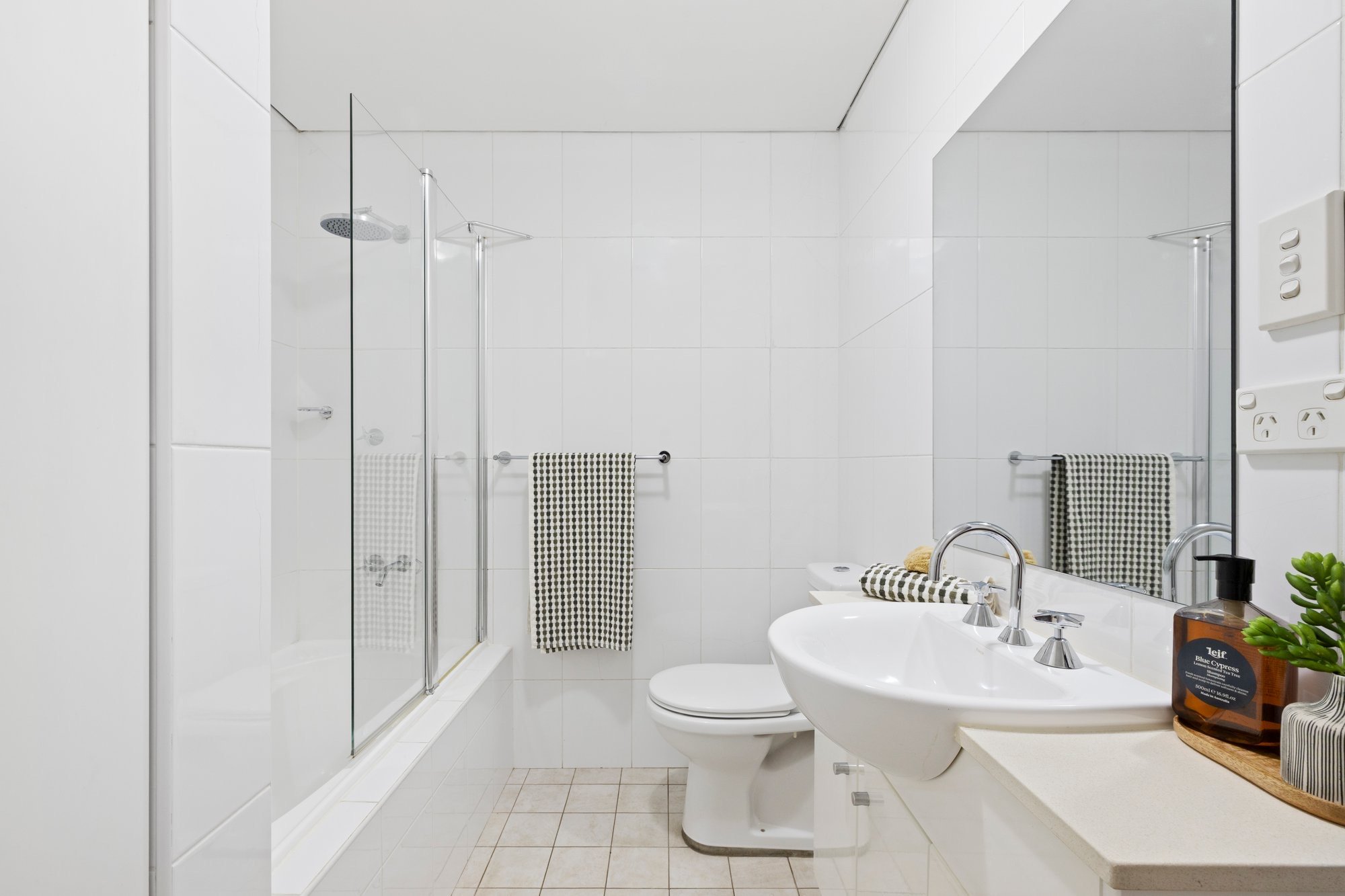 Modern white bathroom with shower area, twin sinks with striped towels, toilet, mirror, and decorative items.
