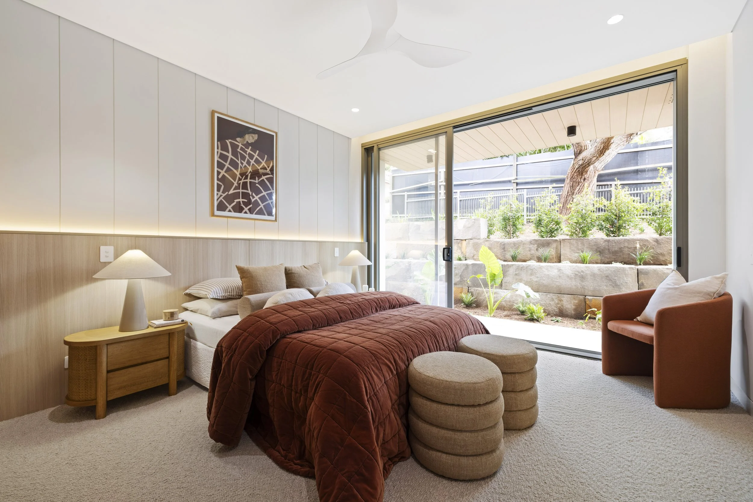 Bedroom with large sliding glass door opening to garden, beige carpet, wooden side table with lamp, bed with rust-colored quilt, upholstered armchair, and two round beige ottomans.