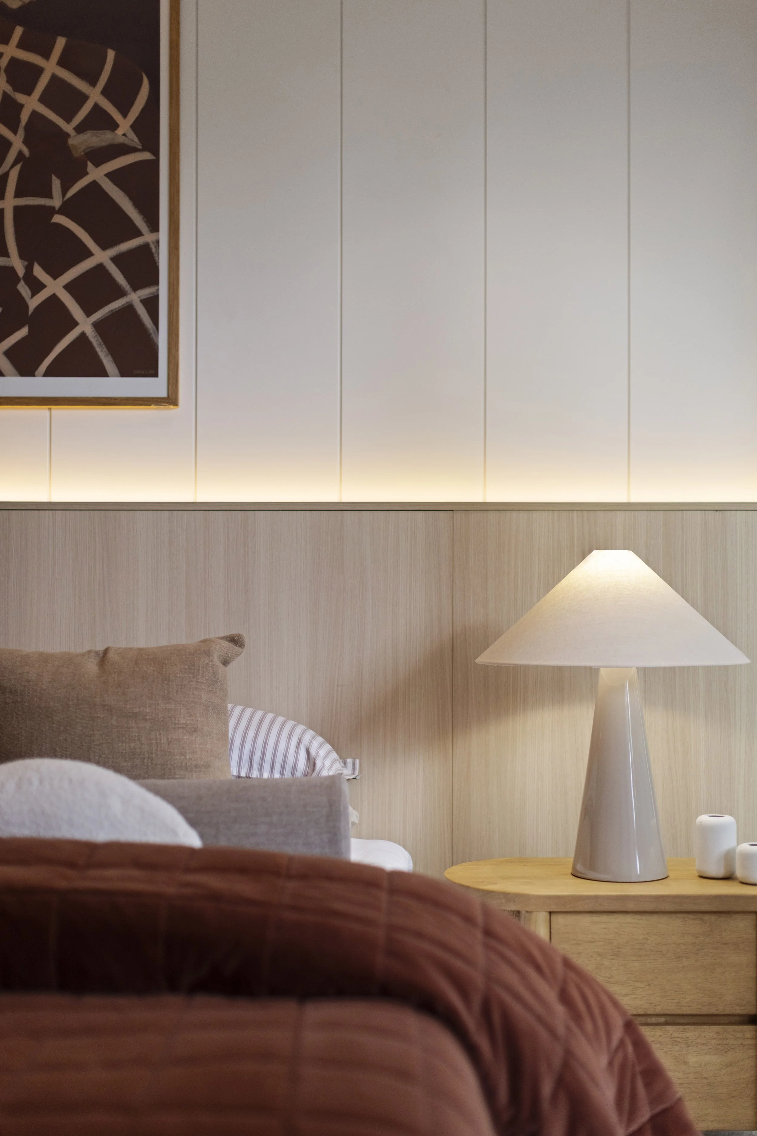 A cozy bedroom corner with a bed, decorative pillows, a wooden bedside table, a modern white bedside lamp, and framed artwork on a light-colored paneled wall.