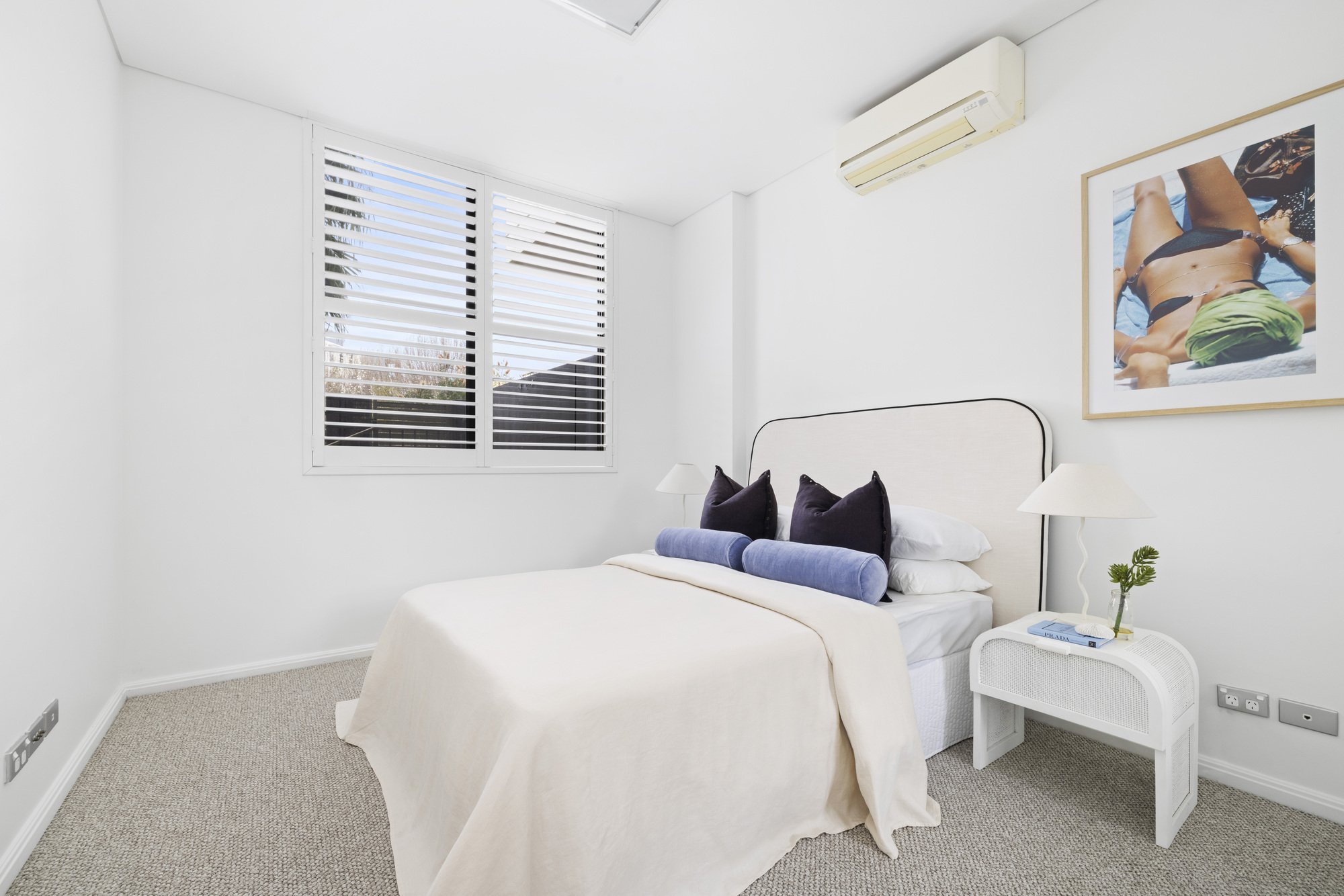 A bright bedroom with a white bed, navy and purple pillows, a small white nightstand with a plant, book, and lamp, a window with white blinds, and a framed photo of a person sunbathing on the wall.