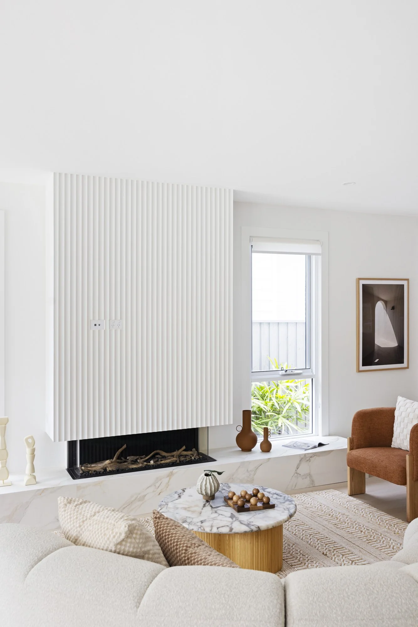 Modern living room with white walls, a fireplace with a textured white surround, and a marble hearth. There is a window with a view of greenery, decorated with brown vases and books. In the foreground, a cream-colored sectional sofa with textured pil