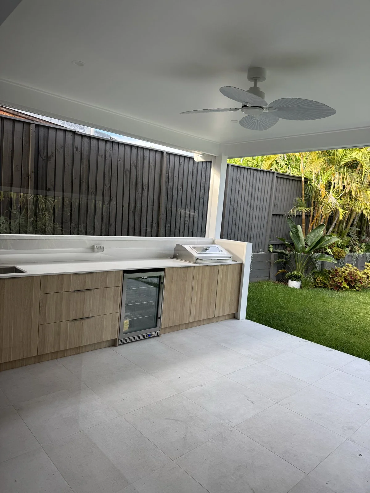 Outdoor kitchen area with wooden cabinetry, mini fridge, countertop, ceiling fan with large leaves-shaped blades, gray tiled floor, black privacy fence, and lush green plants and trees in the background.