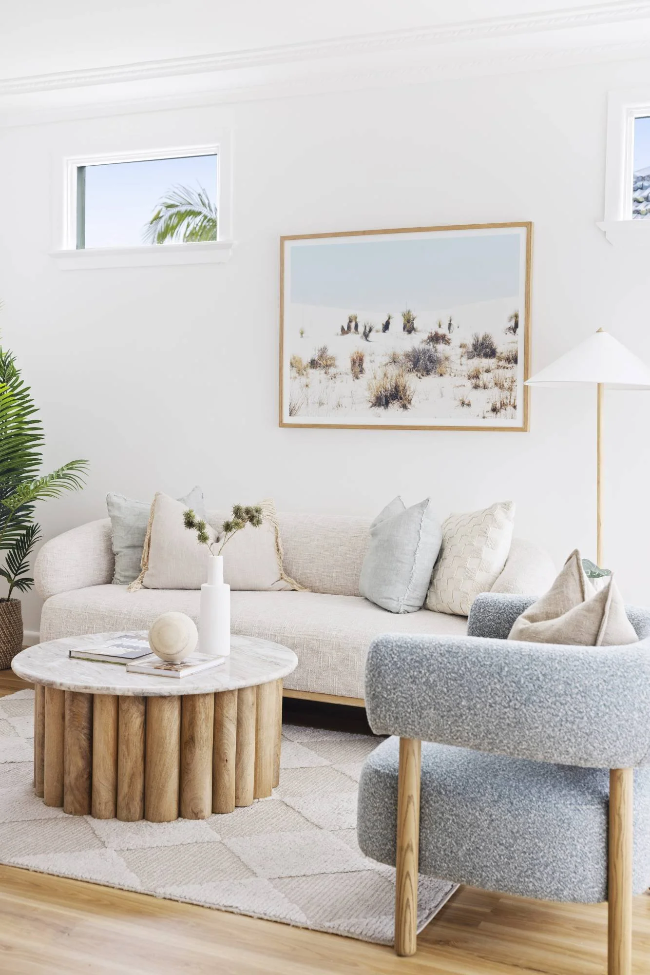 Living room with beige sofa, gray armchair, white side table, and artwork of desert landscape on white wall, with potted plant and windows.