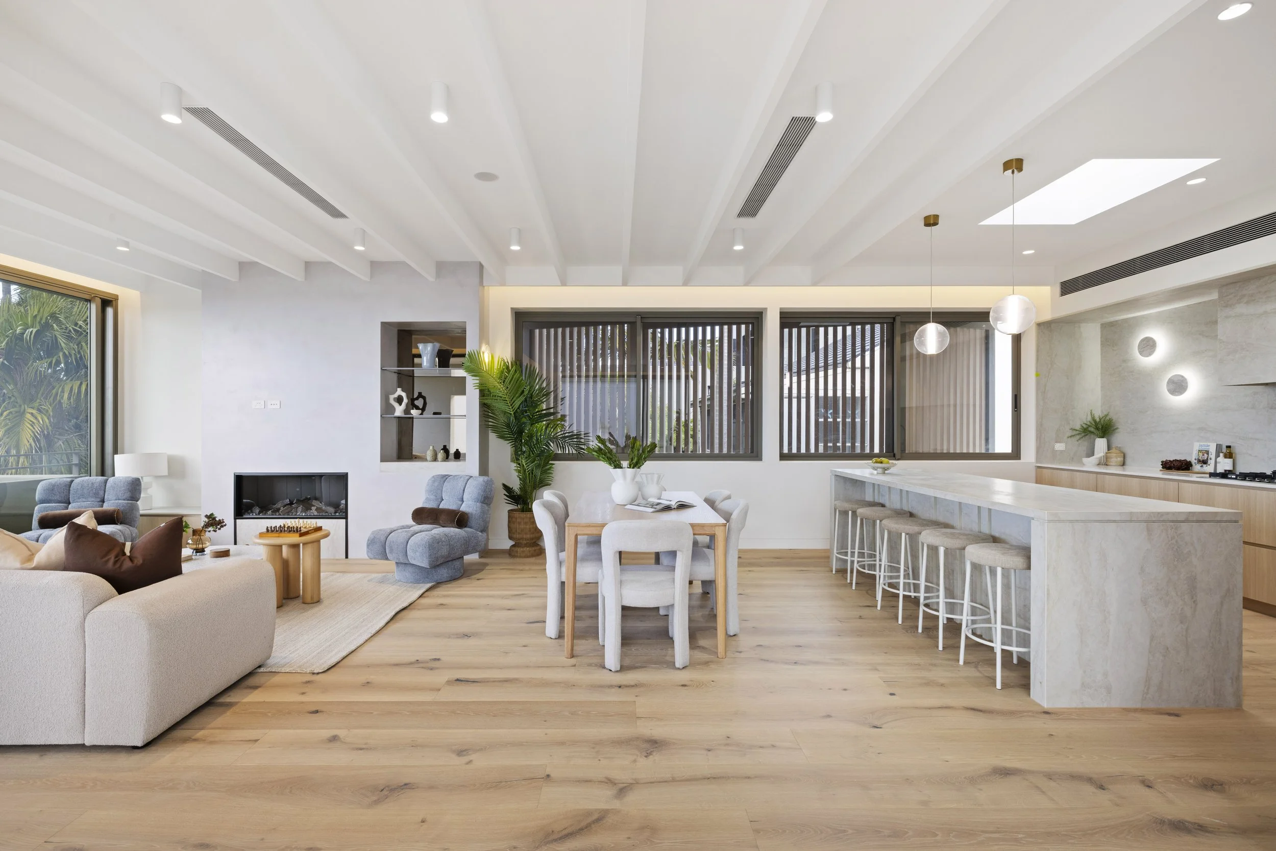 Modern open-concept living room and kitchen with white walls and ceiling, wood flooring, multiple windows, a fireplace, gray sofa, armchairs, a dining table with white chairs, a kitchen island with bar stools, pendant lights, and decorative plants.