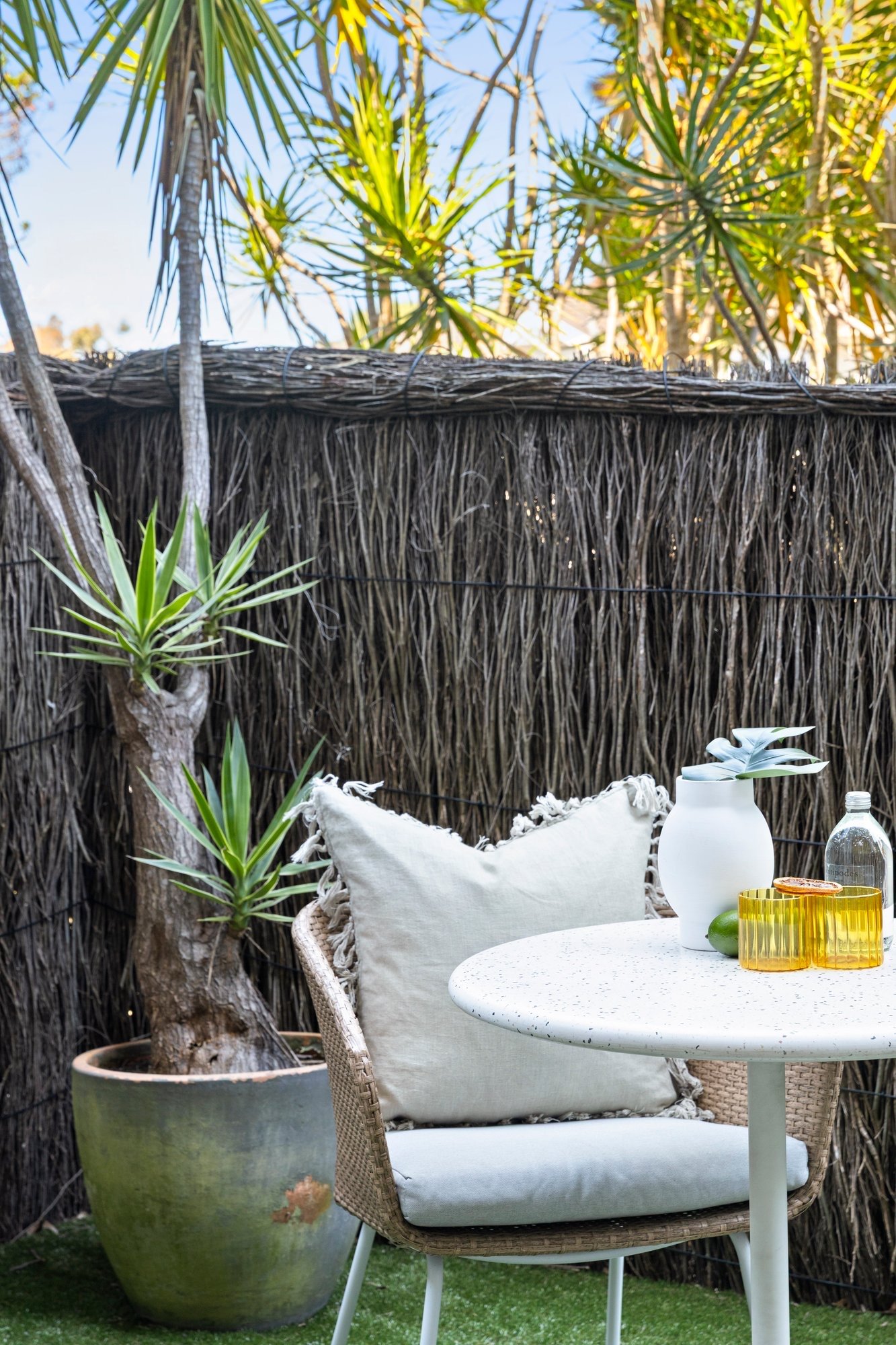 An outdoor patio scene with a wicker chair with a cream cushion and a beige pillow, a white round table with decorative objects including a white vase, a green fruit, yellow glass candle holders, and a glass water bottle, surrounded by a tall potted 