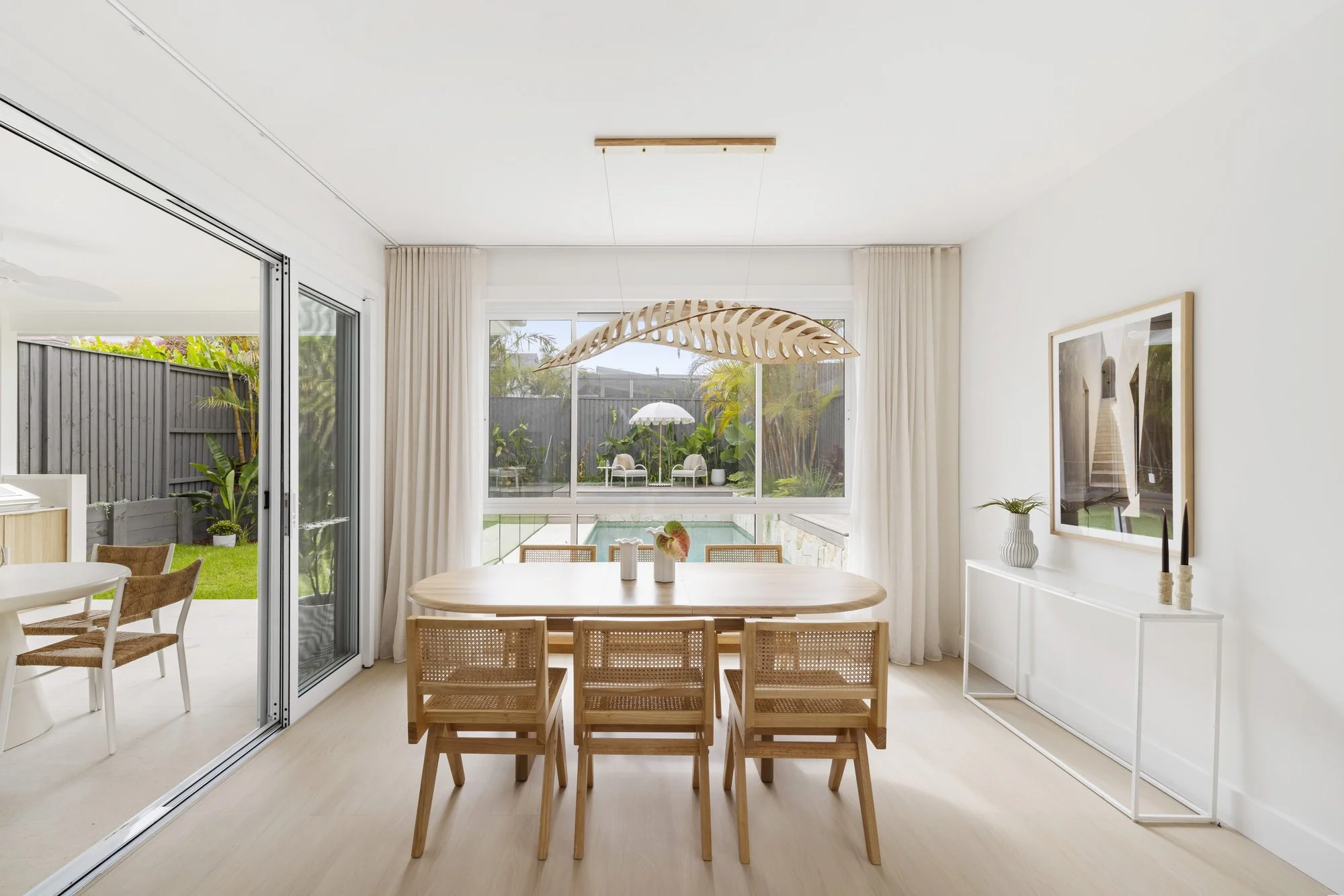 A bright dining room with a wooden table and chairs, large windows with beige curtains, and a view of a backyard with outdoor furniture, greenery, and a swimming pool.