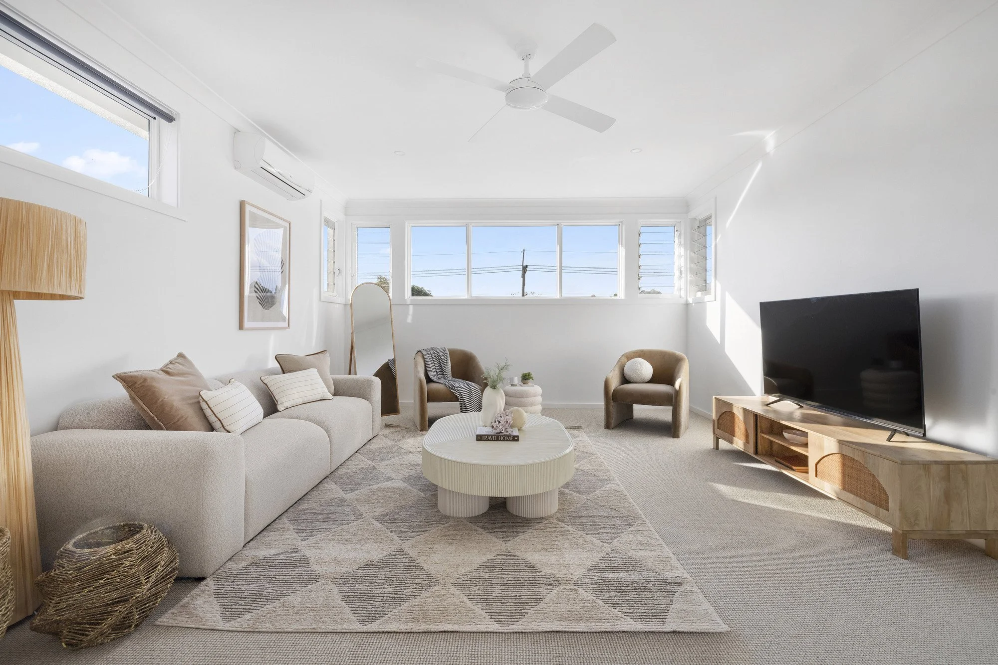 Bright living room with white walls and furniture, beige sofa with pillows, round coffee table, armchair, TV on wooden stand, large windows letting in natural light, ceiling fan, and decorative items.