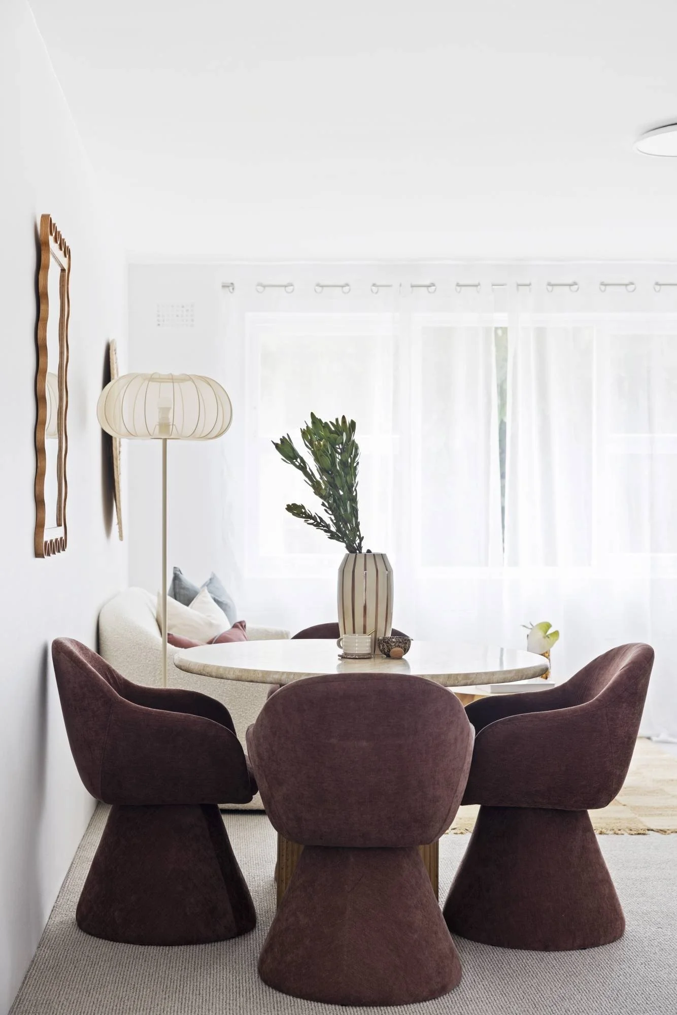 A dining room with dark purple velvet chairs around a round marble table, a large striped vase with green foliage, and a white interior with sheer curtains and wall decor.