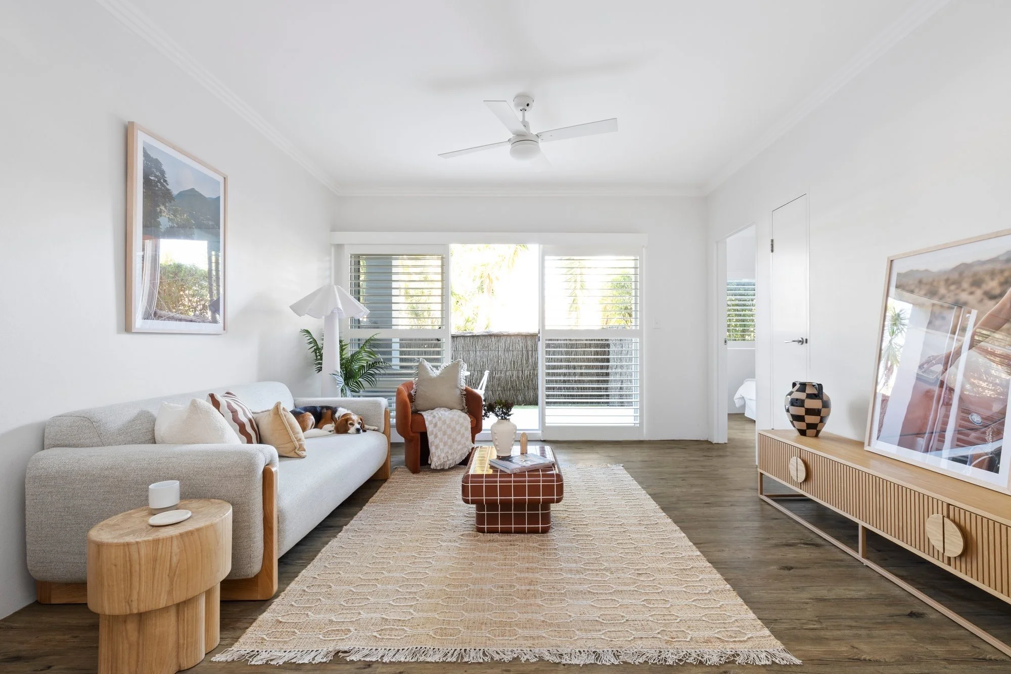 A bright dining room with a wooden table and chairs, large windows with beige curtains, and a view of a backyard with outdoor furniture, greenery, and a swimming pool.