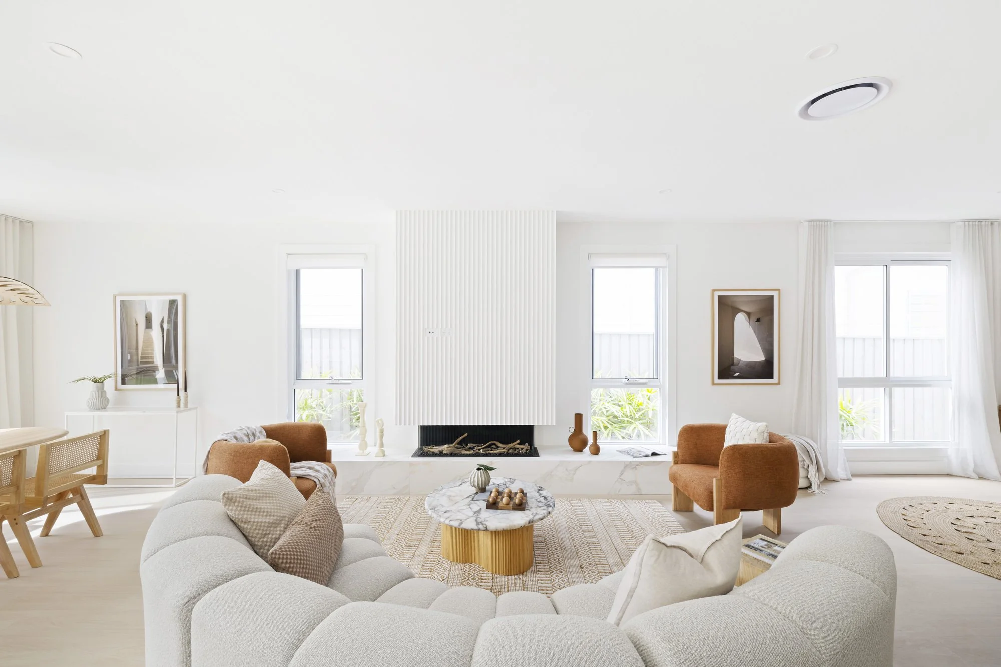 Bright living room with white walls, large windows, beige and orange furniture, a fireplace, and decorative items such as vases and framed pictures.