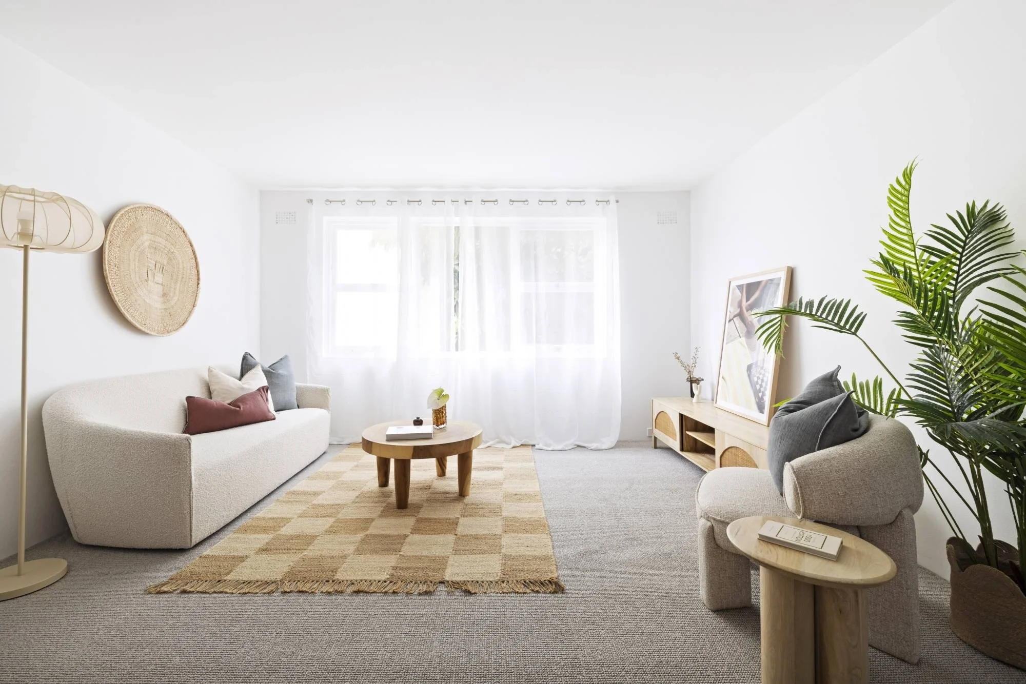 Bright and airy living room with white walls, large window with sheer curtains, beige sofa with pillows, wooden coffee table, large framed artwork, potted plants, and cozy area rug.