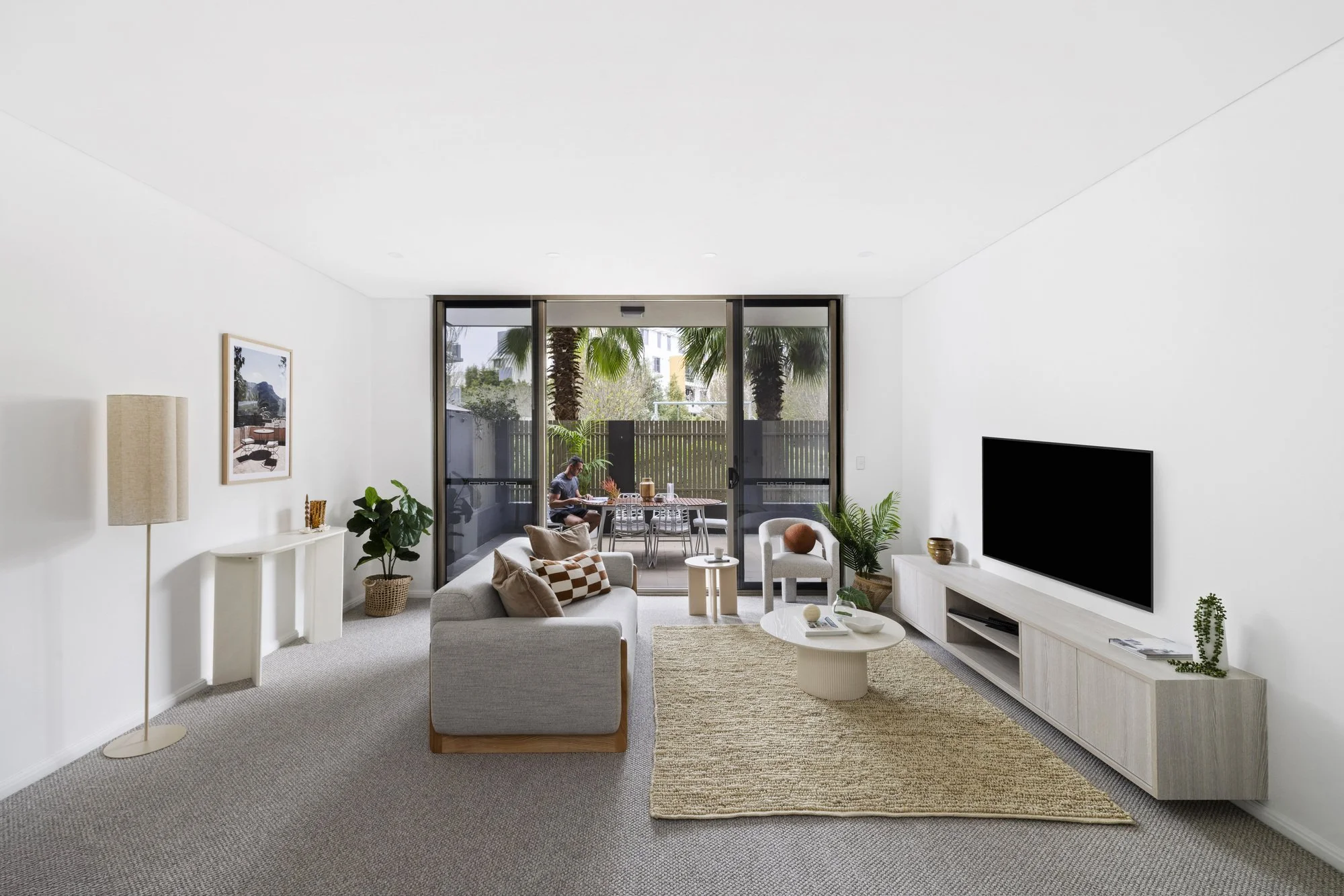 A modern living room with white walls, a grey sofa decorated with checkered and solid pillows, a round white coffee table, and a white media console with a flat-screen TV. There are potted plants and a framed picture on the wall. Large sliding glass 