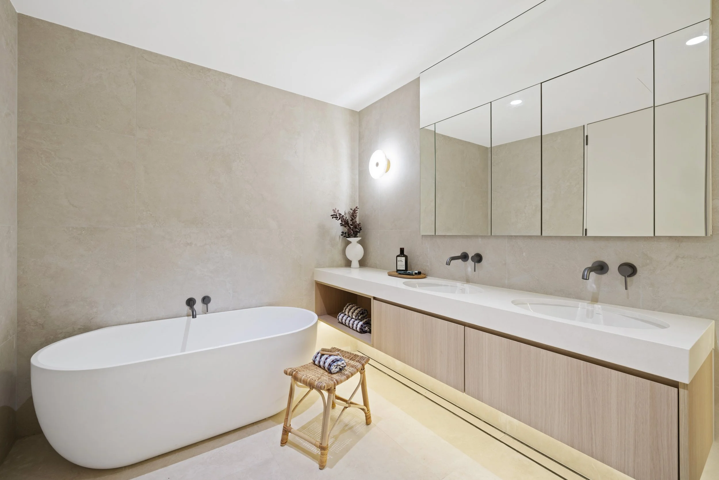 Modern bathroom with beige tile walls and a large mirror. Features a freestanding bathtub, a vanity with two sinks, and decorative items, including a white vase with dried flowers and folded towels.