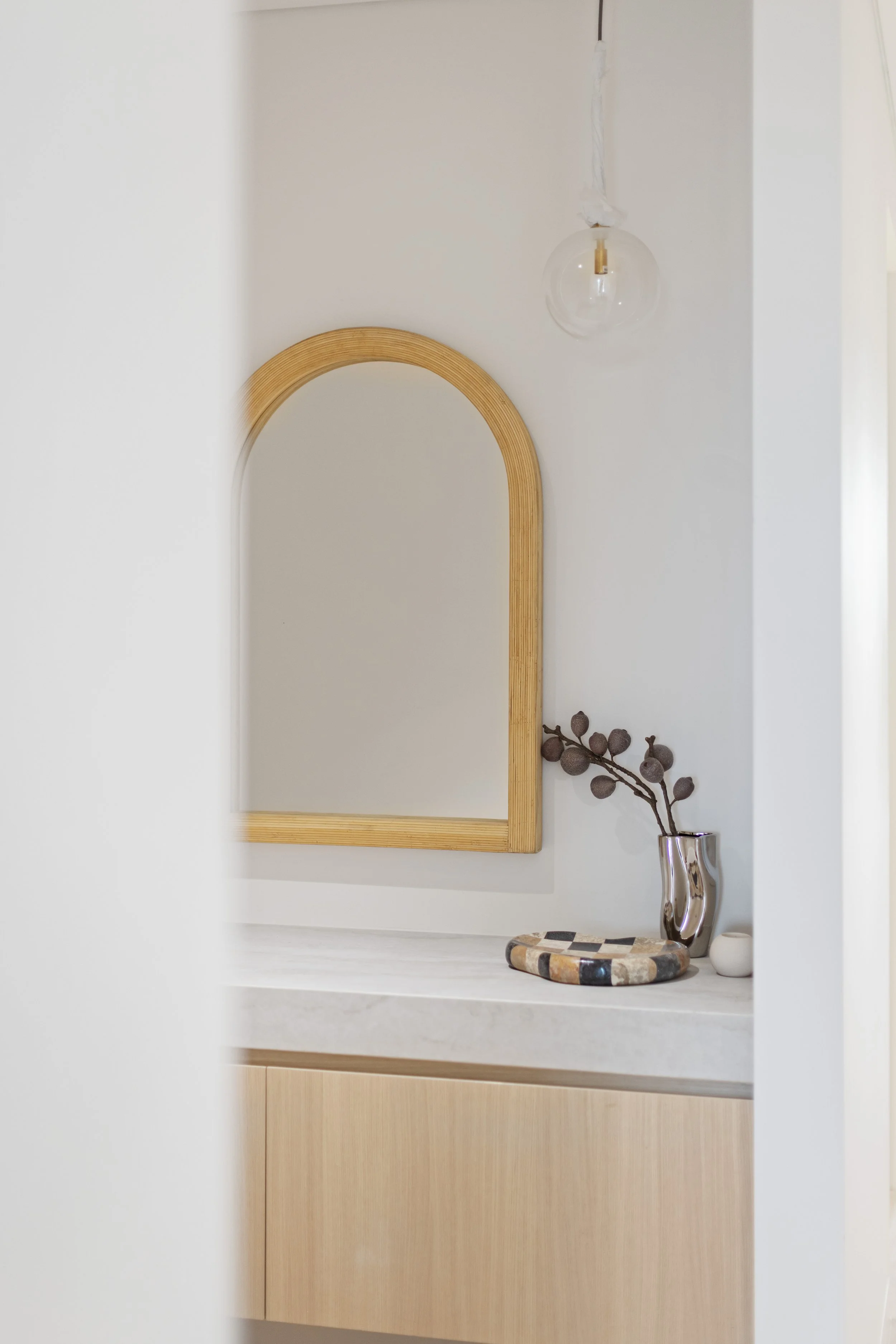 Decorative interior space featuring a wooden arched mirror, a glass hanging light bulb, a metallic vase with dried branch, a patterned tray, and a small white object, all set against a white and wood-finished wall.