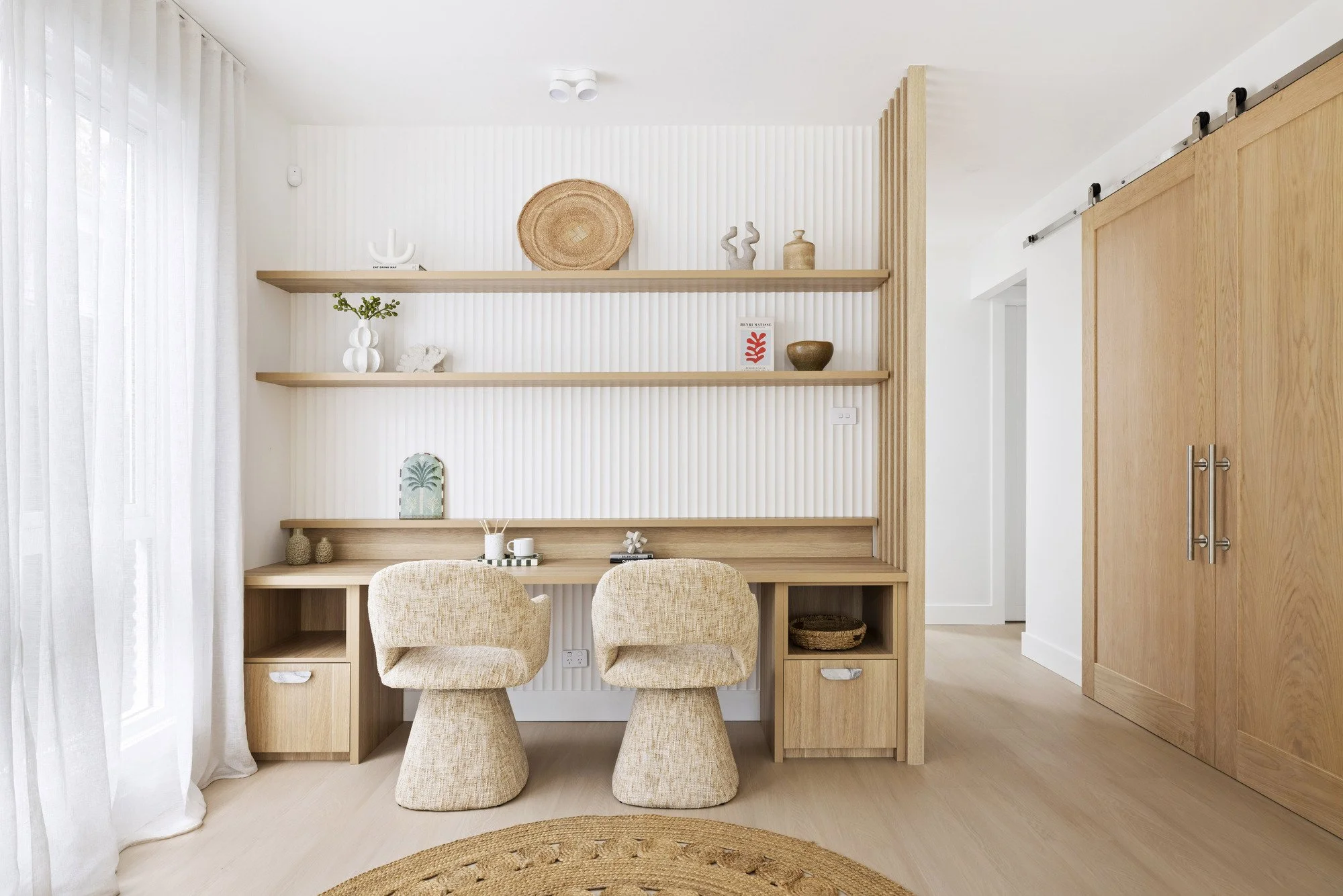 A minimalist home office with a wooden desk, two beige upholstered chairs, open wooden shelves on a textured white wall, decorative items, and a sliding wooden barn door.