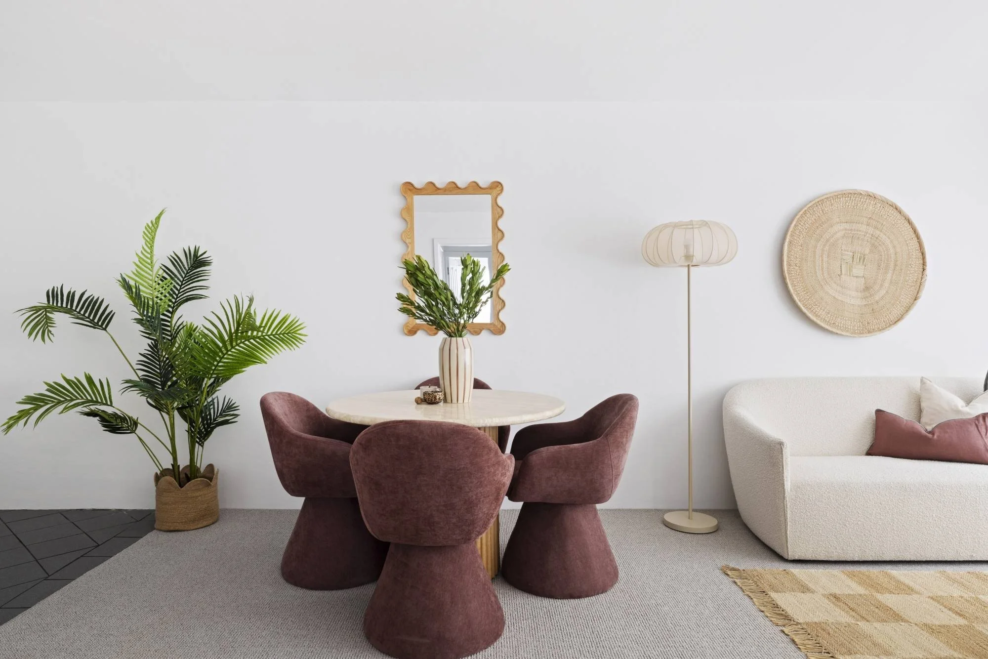 A cozy living room with a round dining table surrounded by four maroon velvet chairs in front of a white wall with a decorative mirror, a floor lamp, and a woven wall hanging. A large green potted plant is placed on the left side of the table, and pa