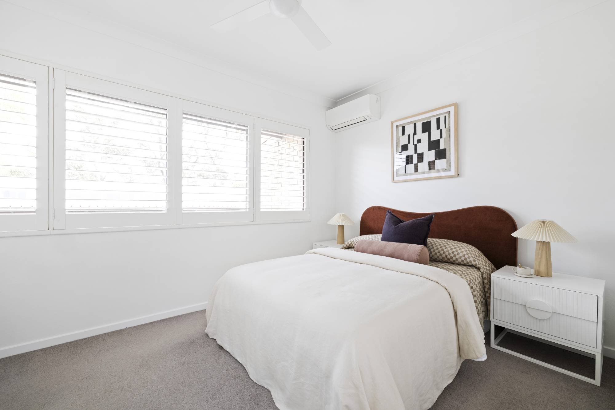 A bedroom with white walls, three windows with horizontal blinds, a bed with white bedding and assorted pillows, two amber bedside lamps, a small white bedside table, and wall art featuring black and white geometric patterns.