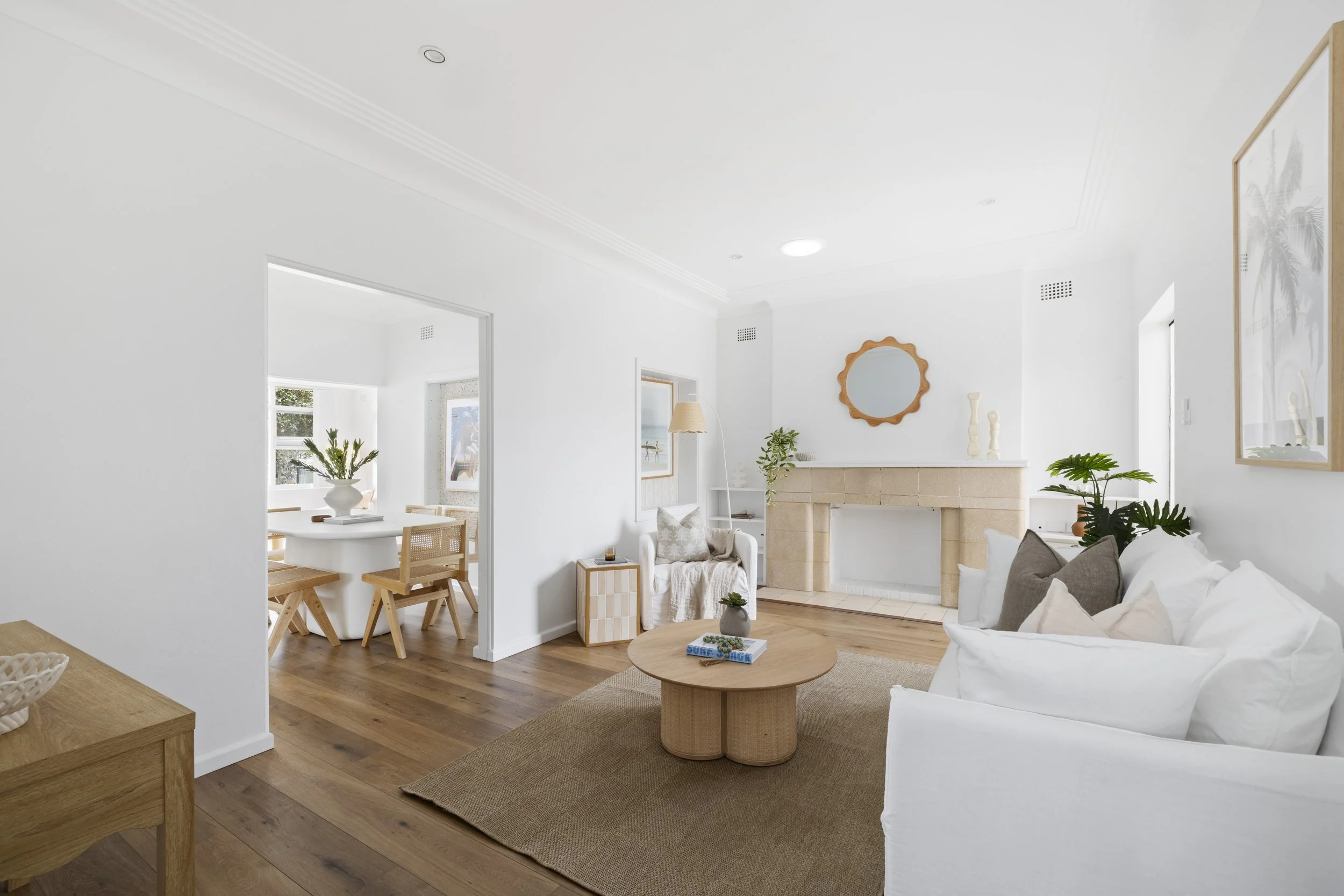 Bright, modern living room with white walls, wooden flooring, and minimalist decor. A fireplace with a mirror above it, a white sofa with throw pillows, a round wooden coffee table with books, and various green plants are visible. Natural light strea