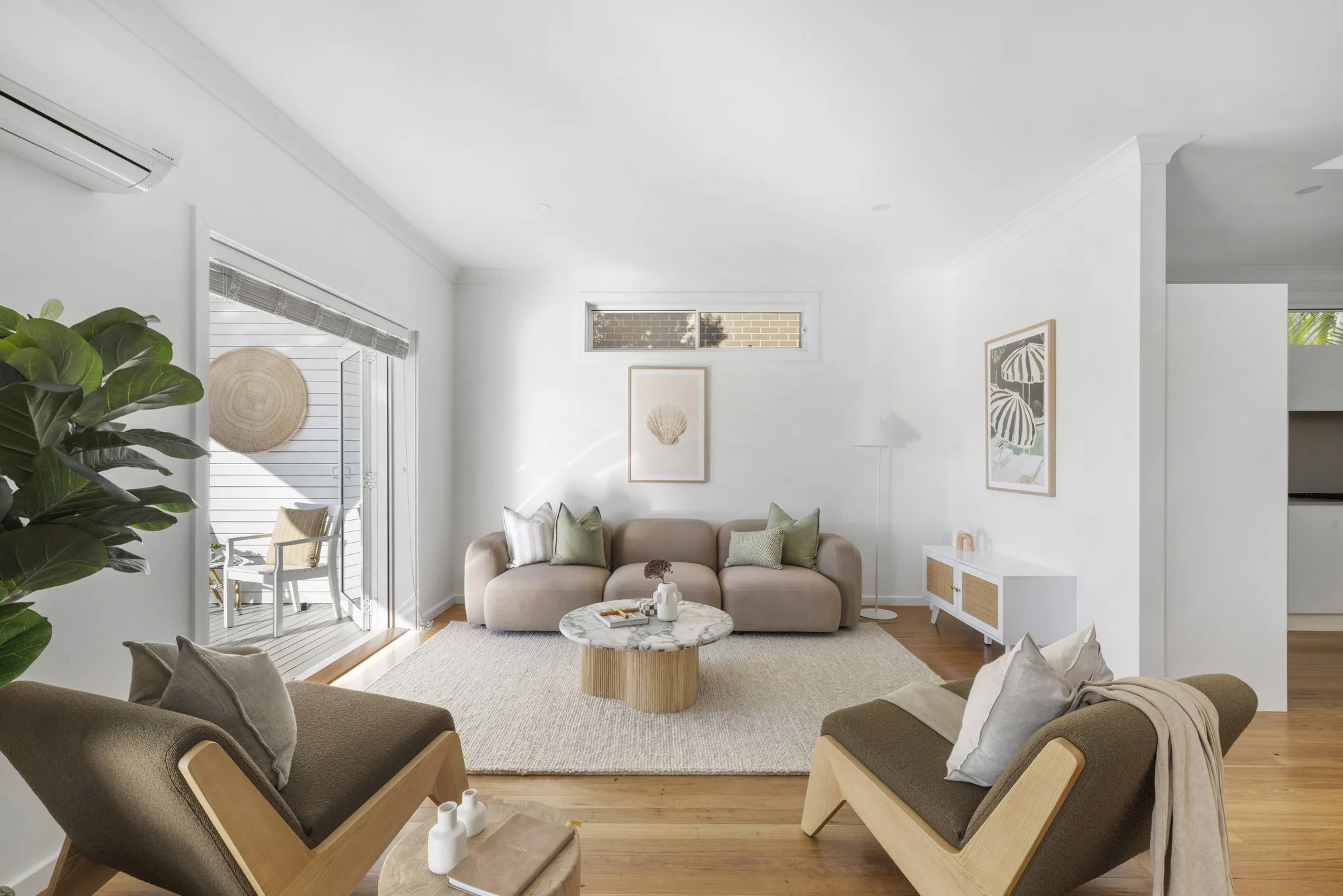 Bright living room with beige sofas, wooden armchairs with gray cushions, round marble coffee table, large green plant, framed artwork on white walls, and sliding glass doors leading to an outdoor patio.