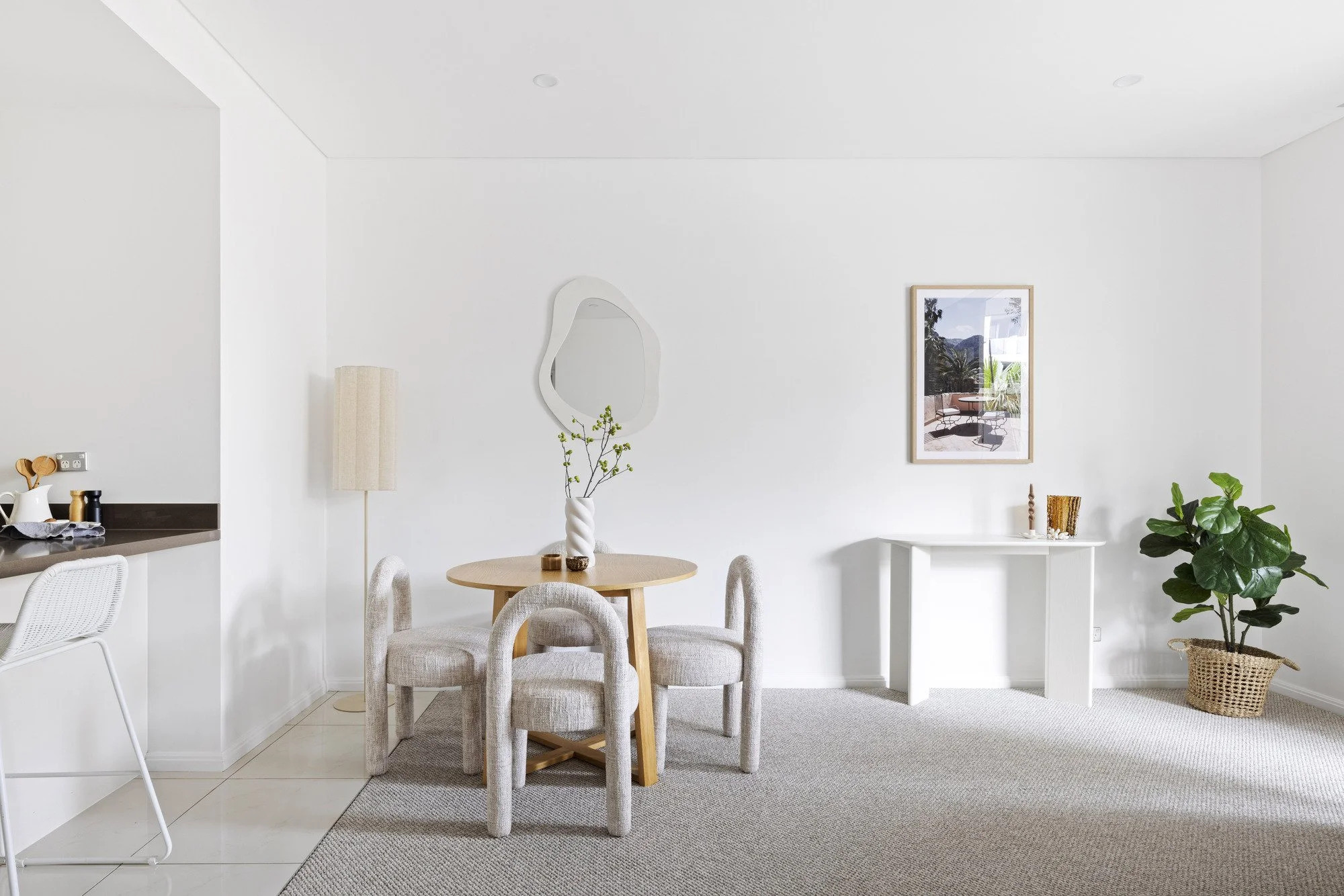 A bright and minimalist dining area with a round wooden table surrounded by four upholstered chairs. The room has white walls, a framed picture of outdoor scenery, a white console table with decorative items, a potted plant, and natural light coming 
