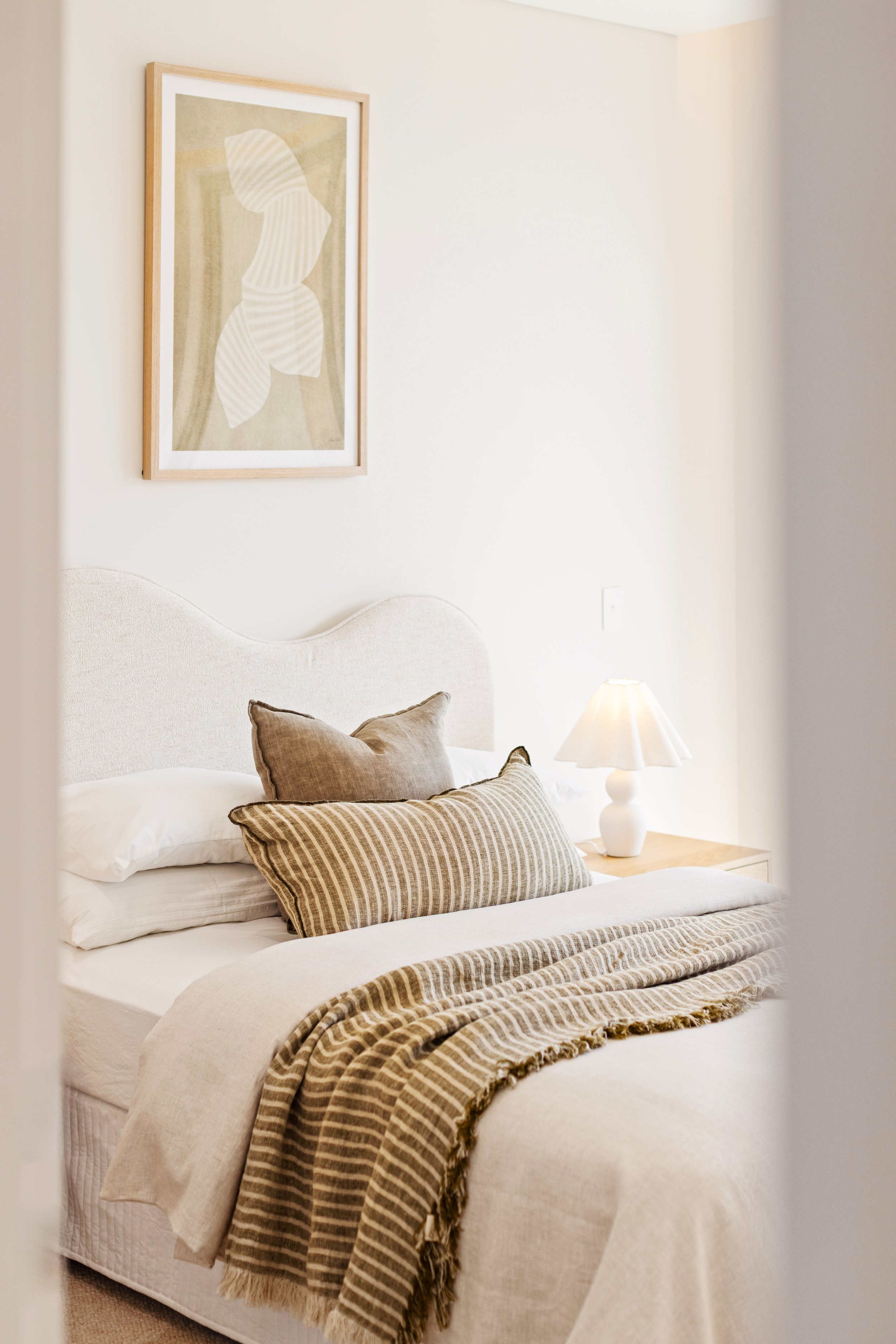 A neatly made bed in a bedroom with a beige headboard, two pillows, a striped throw blanket, a table lamp, and a framed abstract art piece on the wall.