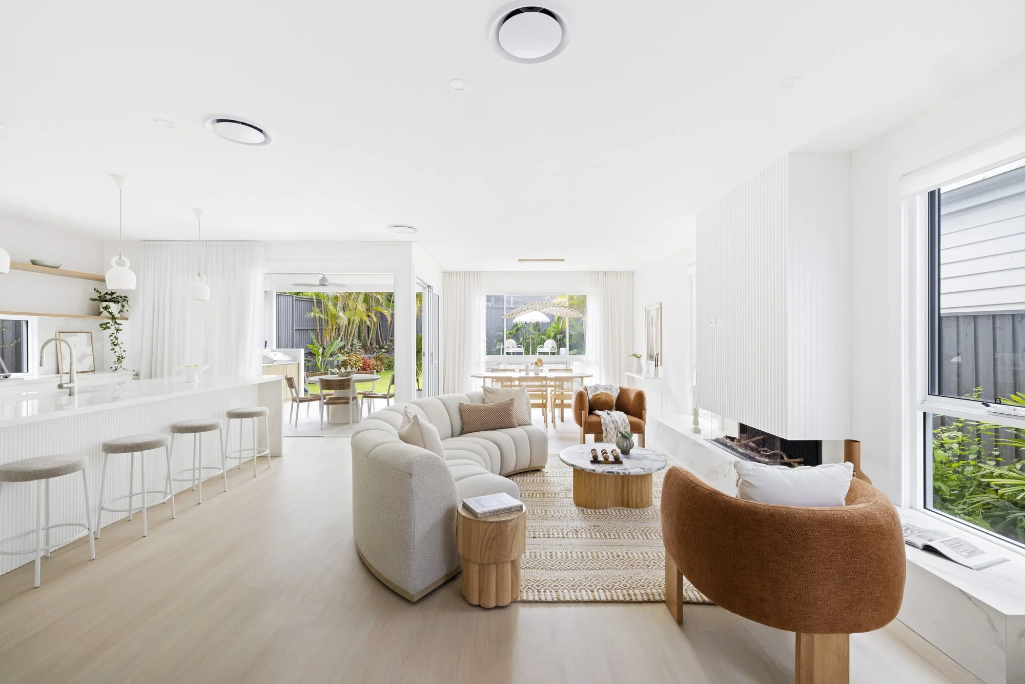 Bright open-concept living room and kitchen with white walls, large windows, and outdoor garden view. Features include a white sofa, orange armchair, coffee table, kitchen island with stools, and dining area with outdoor seating.