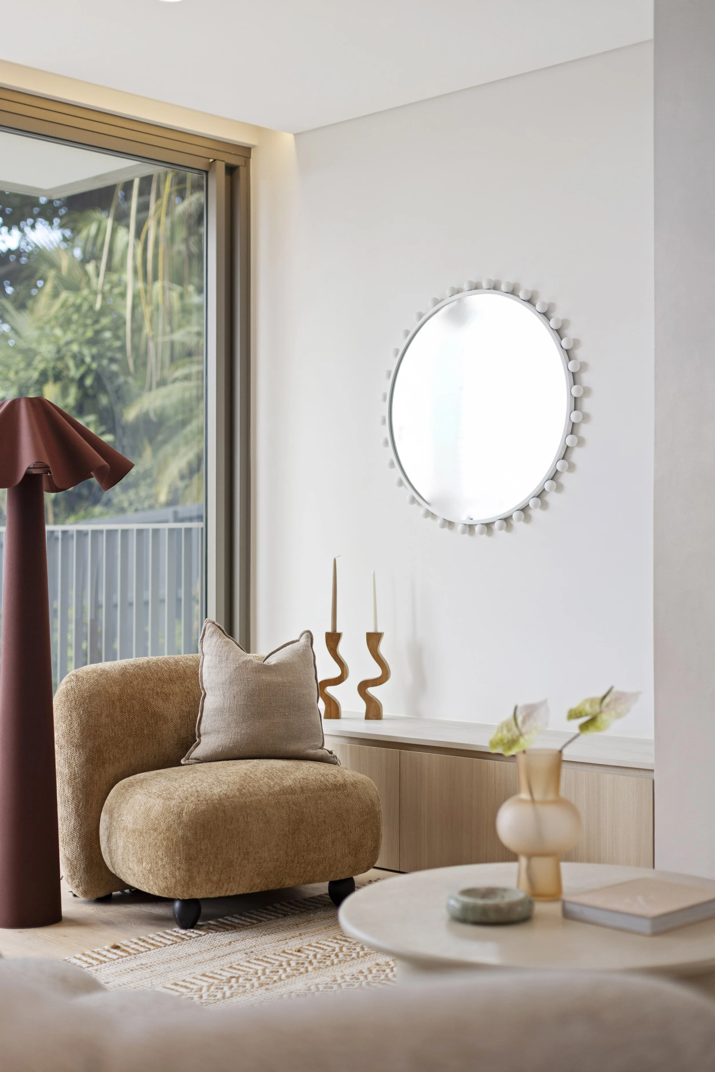 A cozy living room corner with a beige armchair with a pillow, a wooden sideboard with two curvy wooden candle holders, a round mirror with a white frame, a beige vase with a small plant, a window with a sliding glass door showing outdoor greenery, a