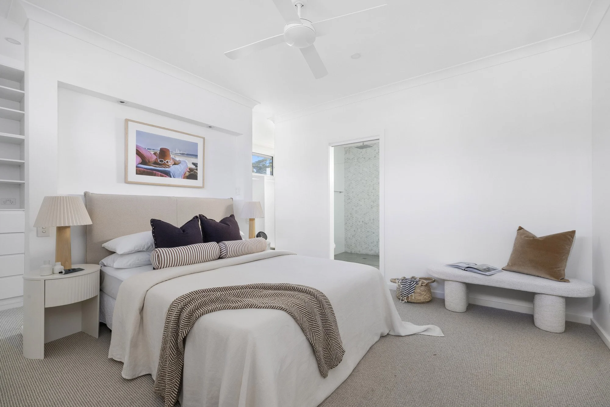 A bright, minimalist bedroom with a white bed, striped bench, and beige headboard. There are two lamps on nightstands, a framed beach photo on the wall, and a window near the ceiling. A doorway leads to a bathroom.