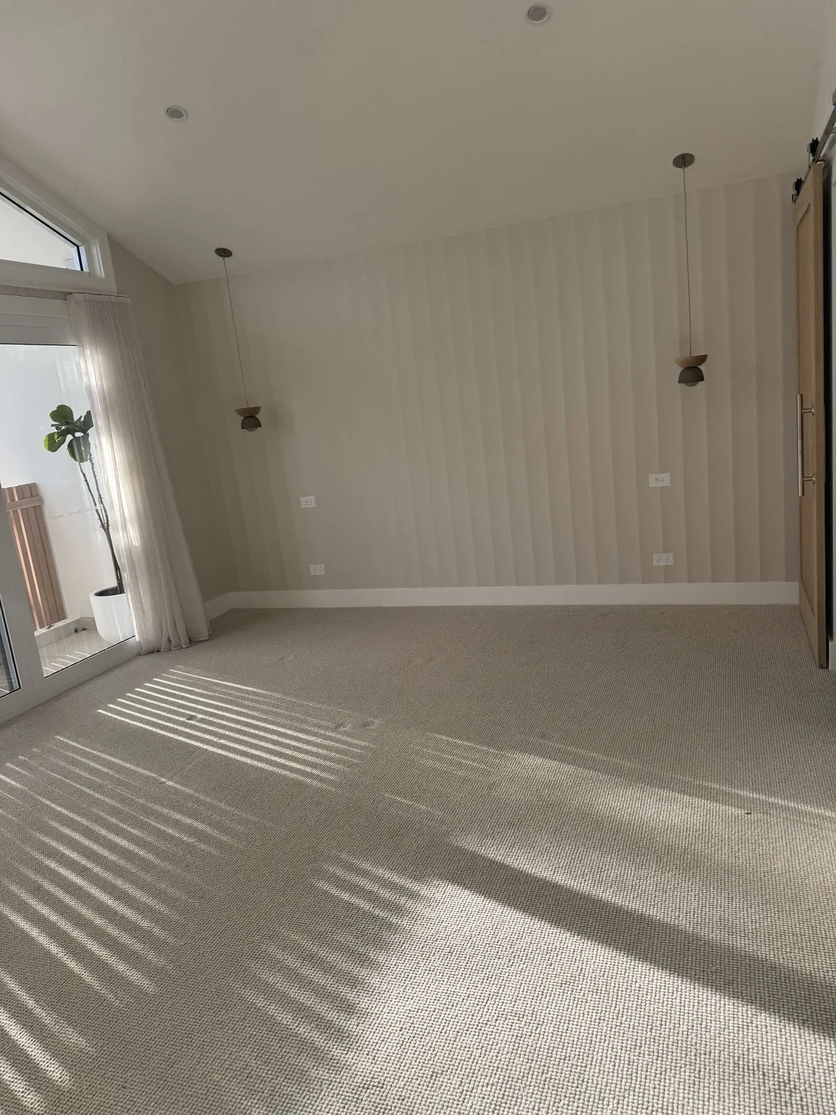 Empty living room with light-colored walls, ceiling pendant lights, a large window with white curtains, and a beige carpet. Sunlight creates striped shadows on the carpet.
