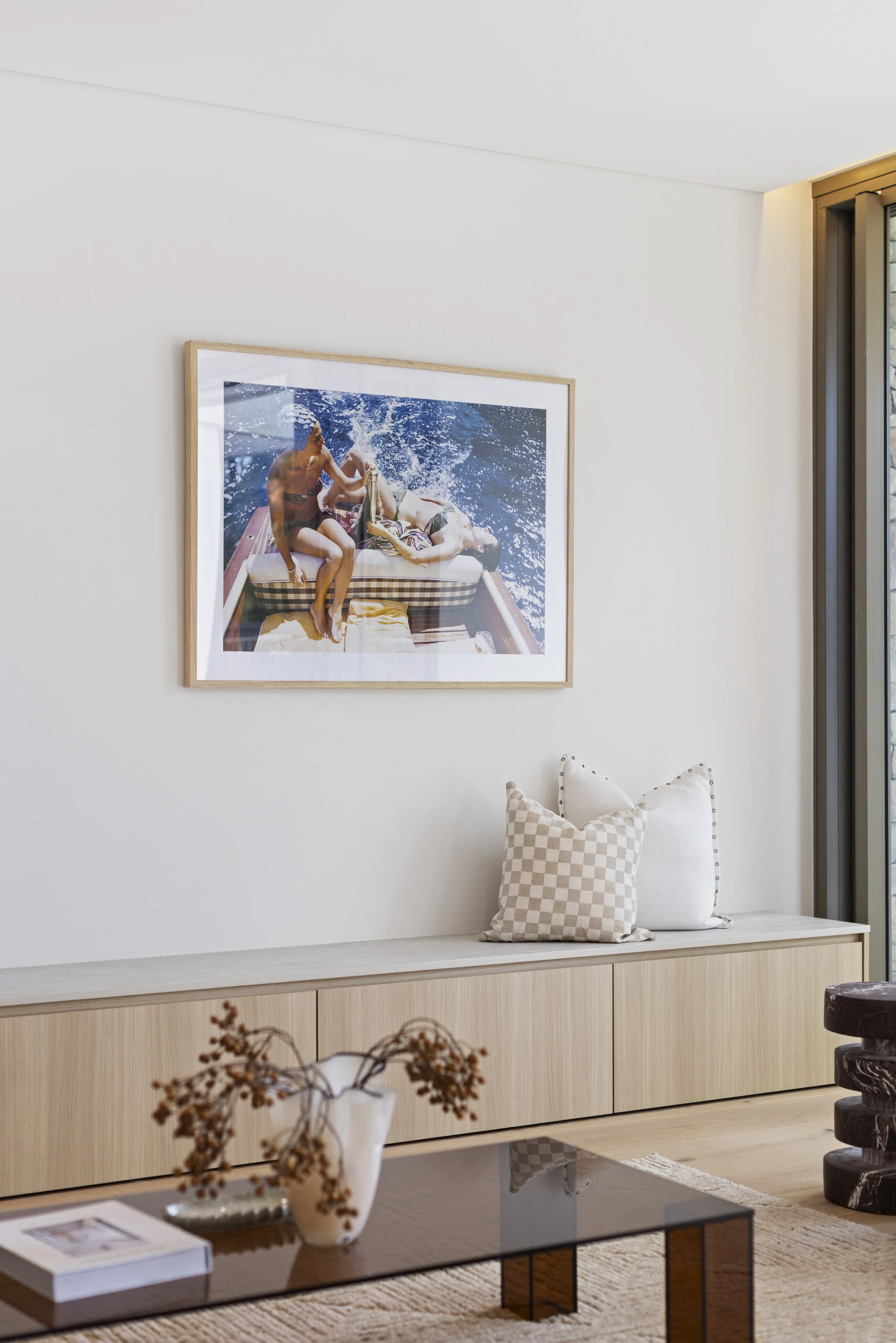 Modern living room with beige sideboard, beige checkered pillows, and a transparent coffee table with a book and a white vase. Framed artwork of two women sunbathing on a boat hangs on the white wall.