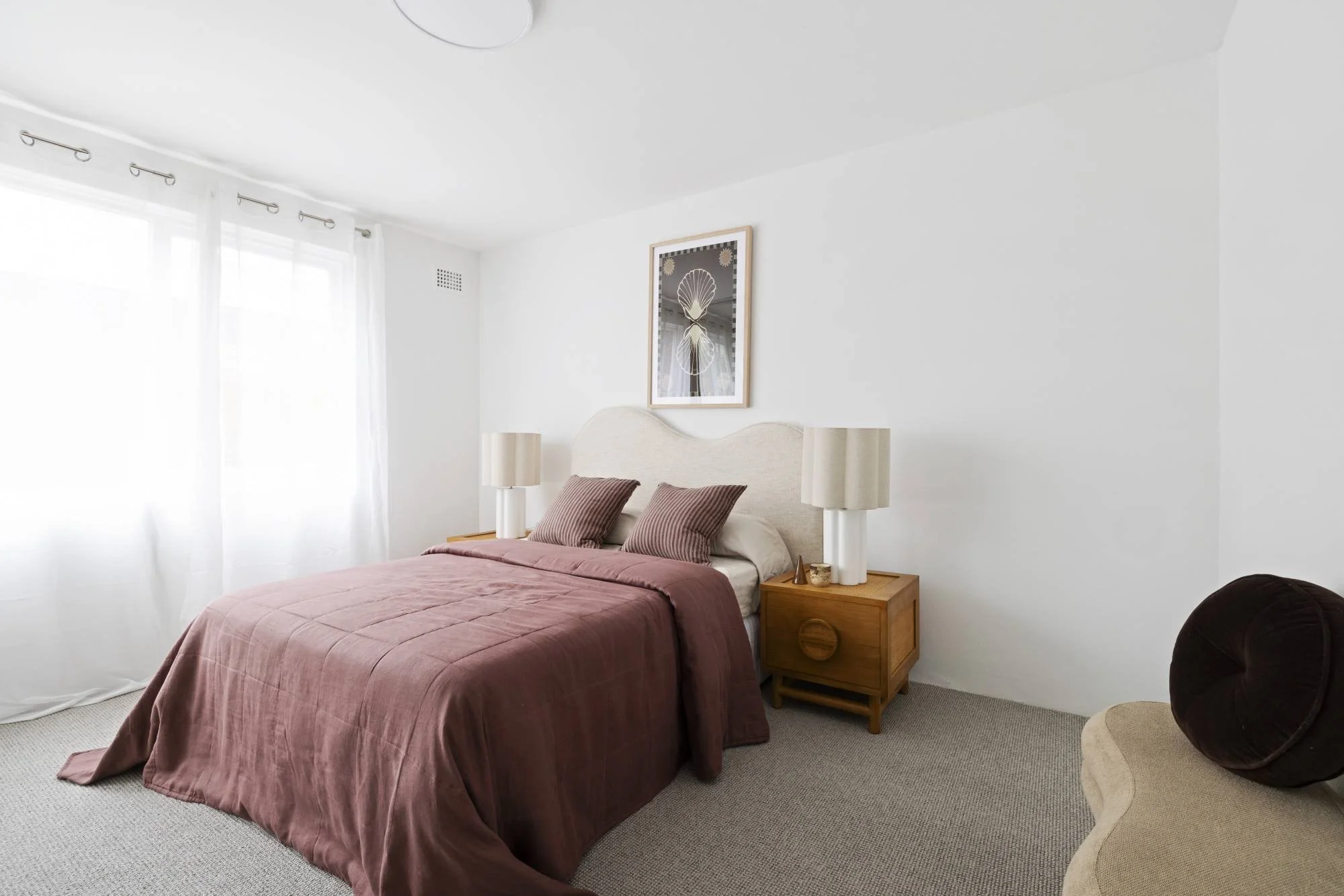 A bedroom with white walls, a bed with a mauve bedspread and matching pillows, two white table lamps on a wooden nightstand, a framed artwork of a seashell above the bed, a window with white curtains, a ceiling light, and a brown round pillow on the 
