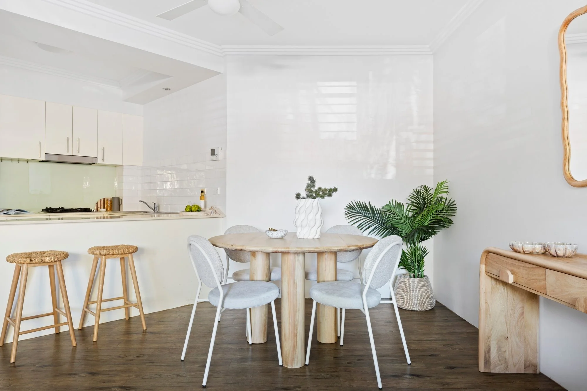 A modern, minimalist kitchen and dining area featuring a round wooden table with four white chairs, a white countertop with cabinets, a small plant in a white textured pot, and decorative objects. The room has white walls, a ceiling fan, and dark woo