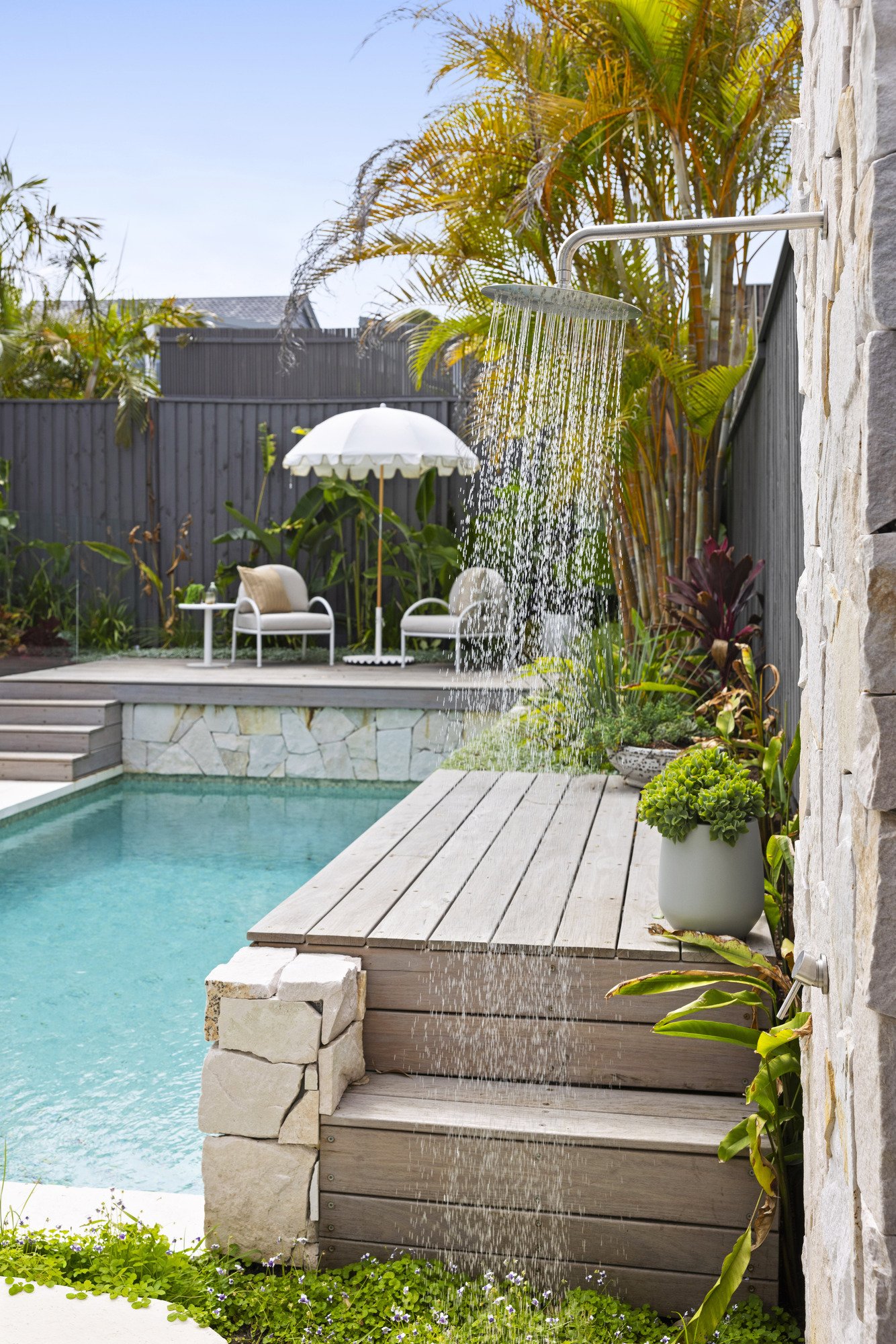 Backyard with swimming pool, wooden deck, lounge chairs, large white patio umbrella, potted plants, waterfall feature from showerhead, tropical plants, and a stone wall on the right.