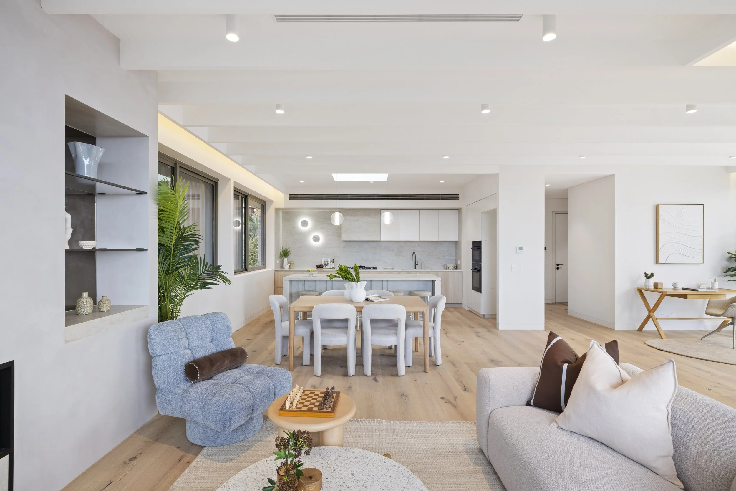 Bright and modern open-concept living room and kitchen in a home with white walls, wooden flooring, and minimalist decor. The space includes a blue tufted armchair, a light wood dining table with chairs, and white sofas. Large windows provide natural