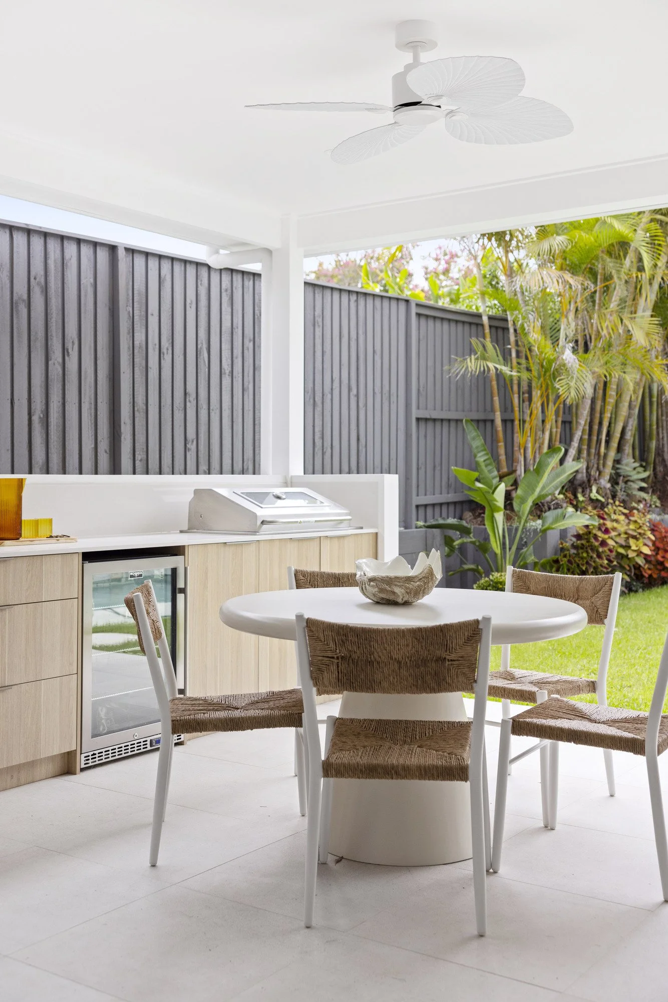 Outdoor patio with a white round table and six chairs with woven seats, a built-in cabinet with a mini fridge, a ceiling fan with leaf-shaped blades, a tall wooden fence, and lush green plants beyond the fence.