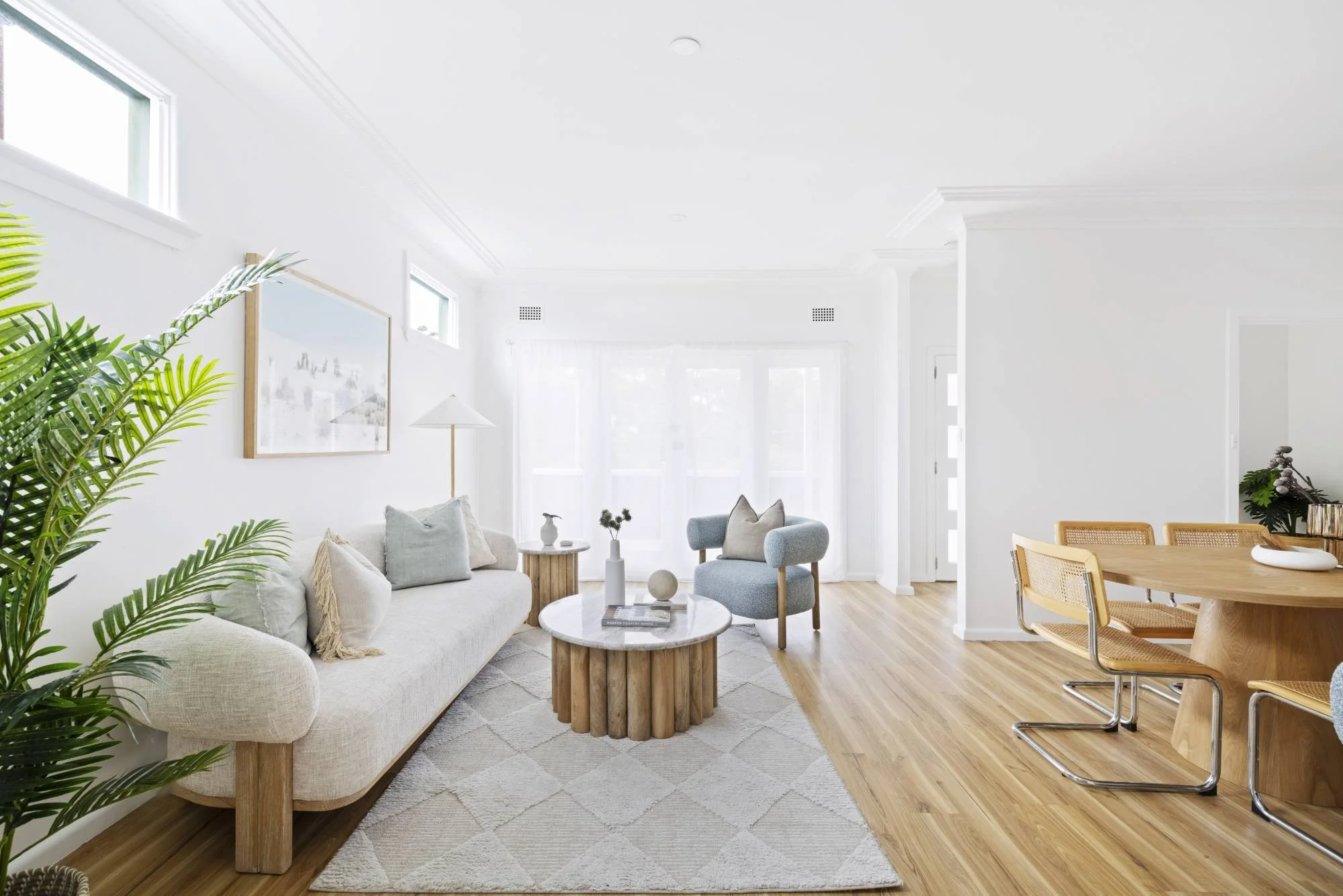 Bright living room with white walls, large windows, green plant, beige sofa with pillows, round wooden coffee table, grey armchair, and dining area with wooden table and yellow chairs.