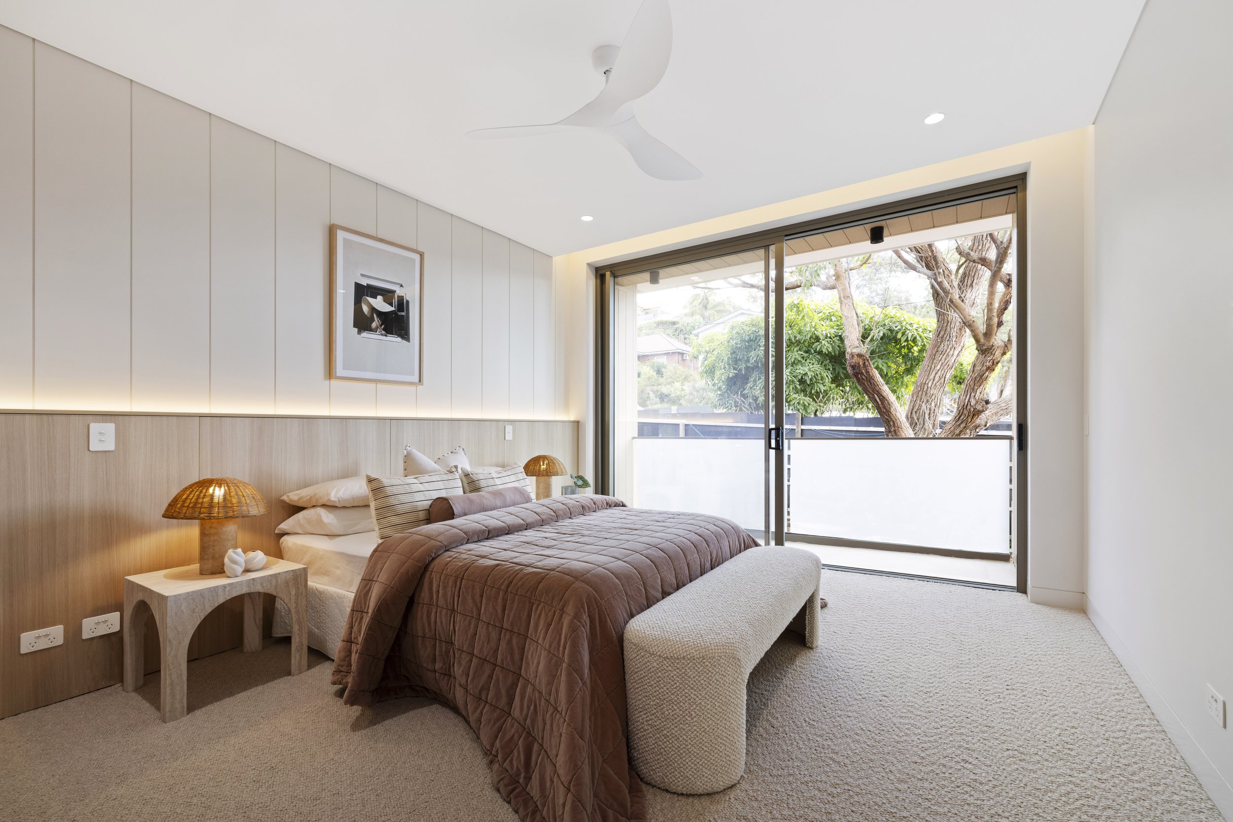 Modern bedroom with a beige upholstered bed with brown bedding, two wicker lamps on side tables, a framed artwork on the wall, and large sliding glass doors opening to a view of trees outside.