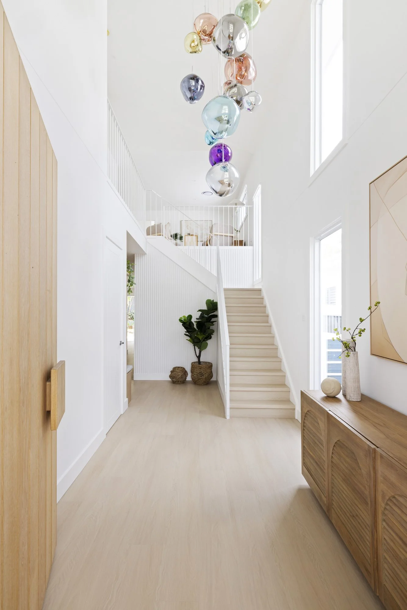 Bright and airy modern home interior with staircase, light wood flooring, white walls, large windows, decorative vases and artwork, and colorful hanging glass art installation.