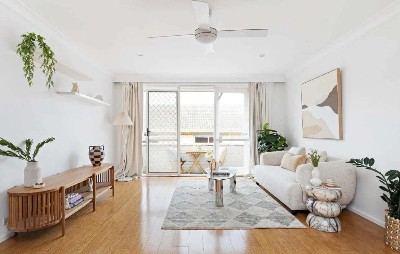 Living room with beige sofa and two armchairs, a round coffee table, a framed picture on the gray wall, a TV on a wood panel wall, and large windows with curtains showing greenery outside.