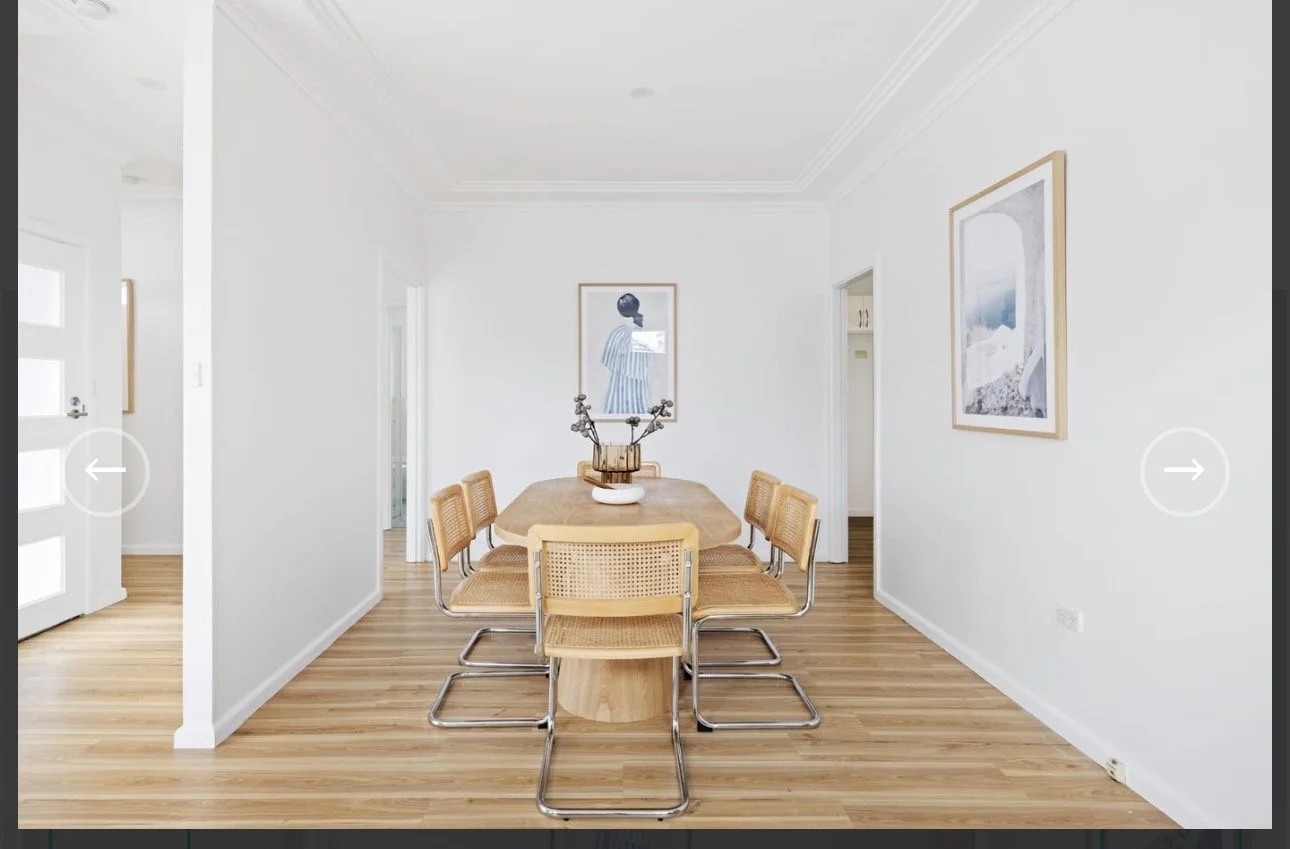 A bright dining room with a wooden table and chairs, large windows with beige curtains, and a view of a backyard with outdoor furniture, greenery, and a swimming pool.
