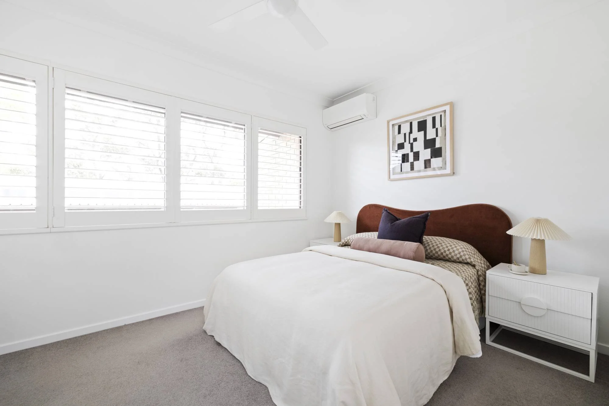 A bright, modern bedroom with white walls and large windows with blinds. The bed has a white duvet, brown and checked pillows, and a curved wooden headboard. There are two beige table lamps on a white nightstand beside the bed. A framed abstract blac