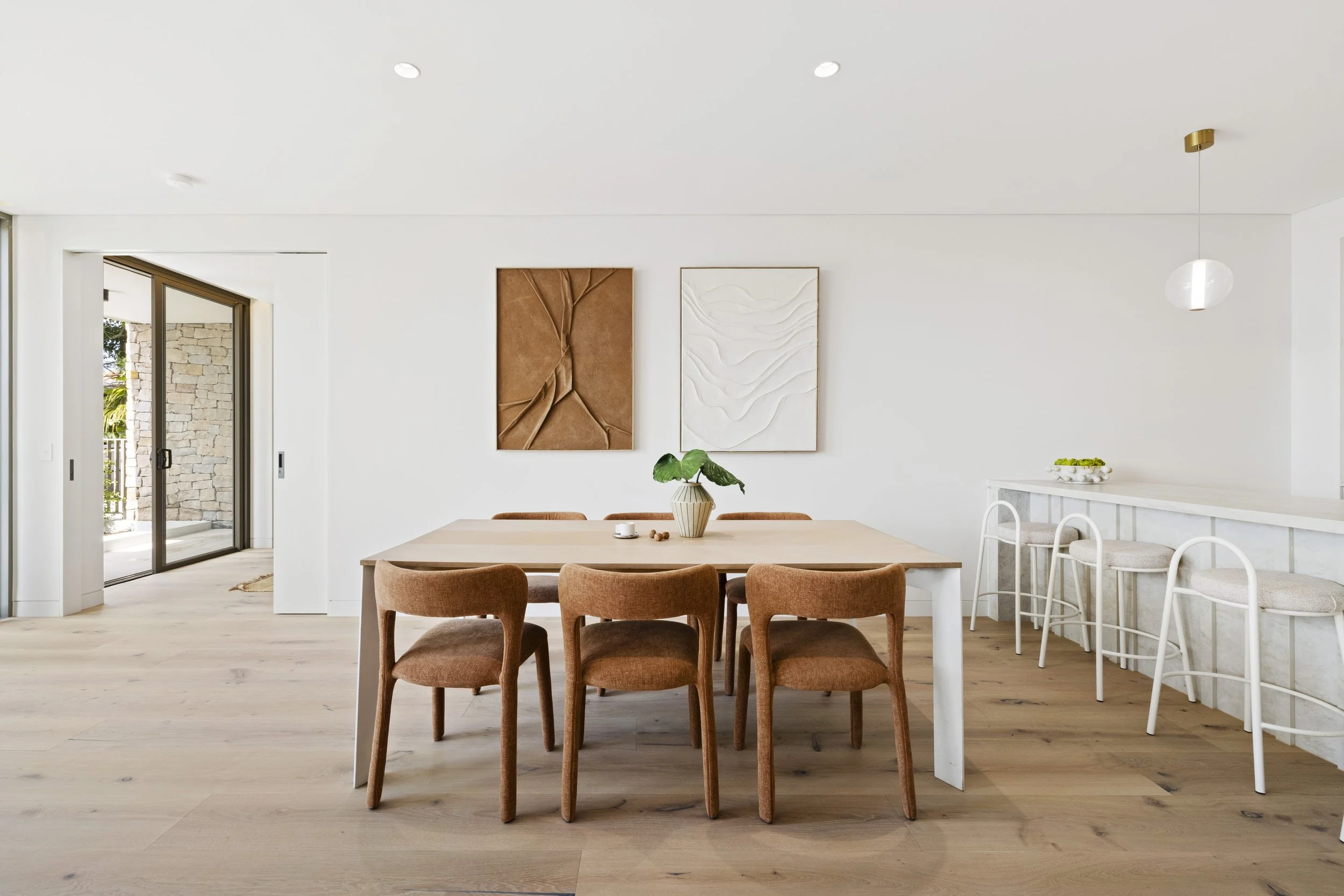 Modern dining room with a wooden table, orange upholstered chairs, and wall art, adjacent to a kitchen counter with barstools and a bowl of lemons on top.
