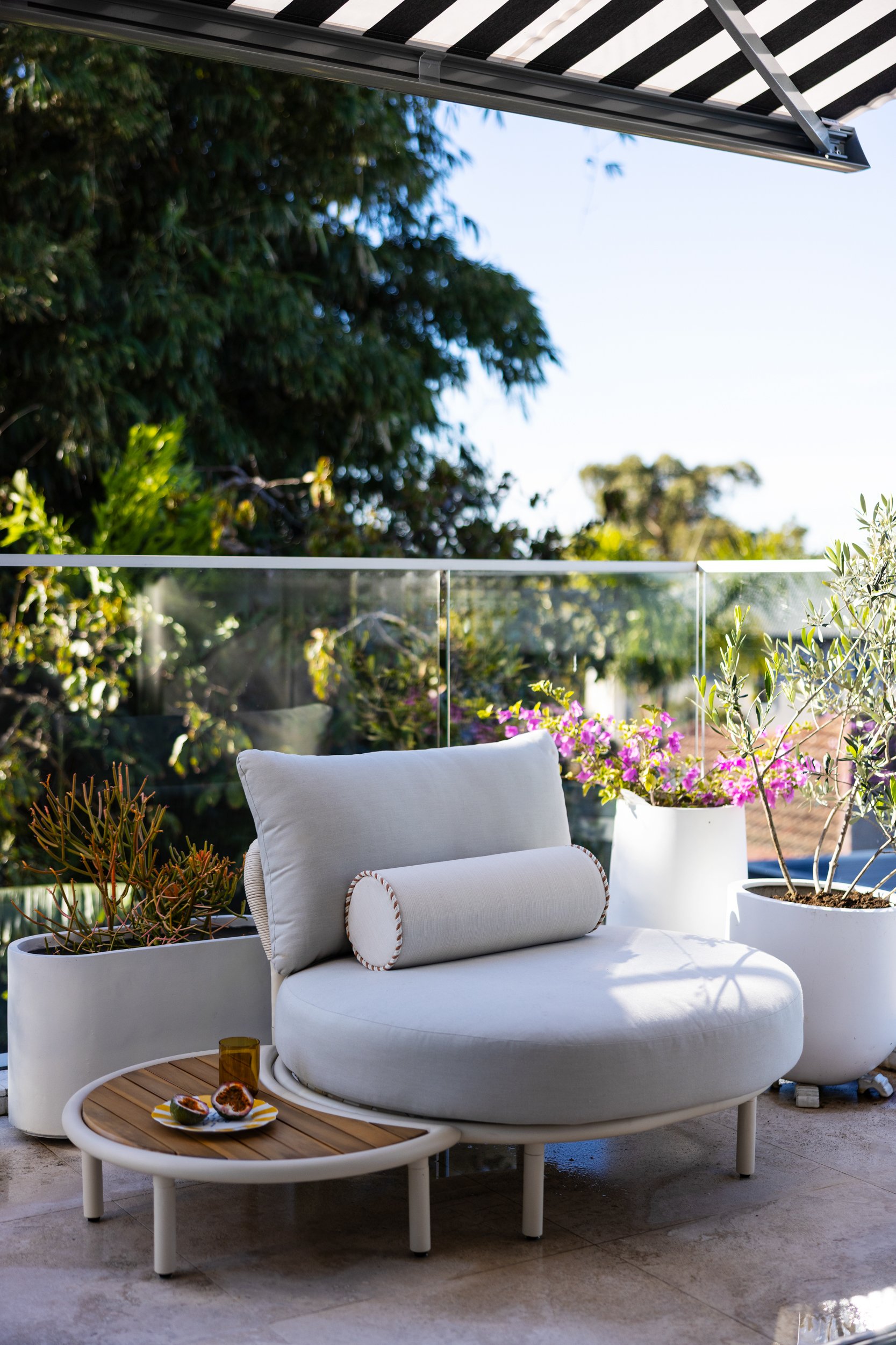 Outdoor balcony with white cushioned lounge chair, surrounded by potted plants and flowers, with a backdrop of green trees and a clear blue sky.