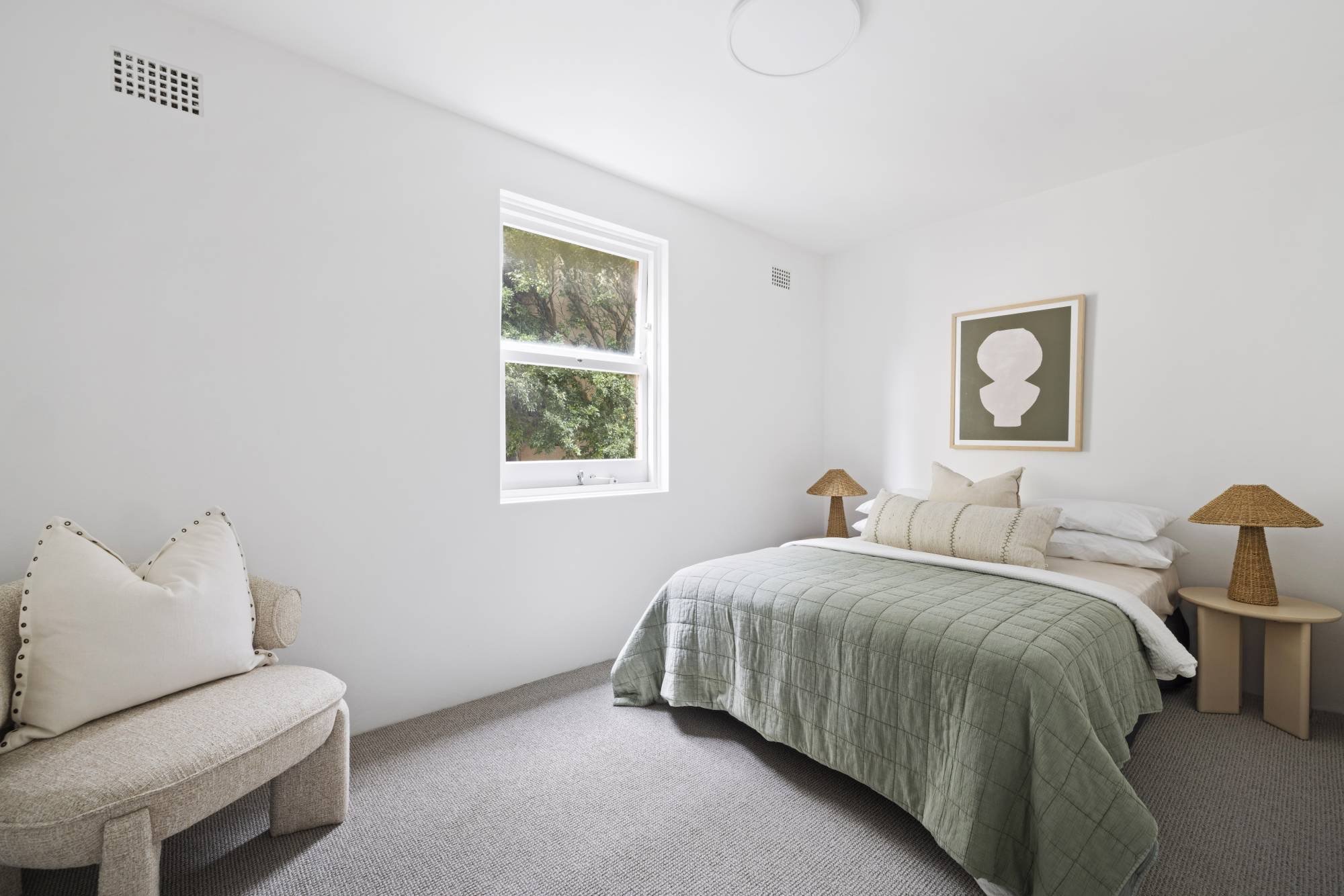 A bedroom with white walls, a window with a tree view, a bed with green quilt, a framed abstract art piece, bedside tables with woven lamps, and a beige chair with pillows.