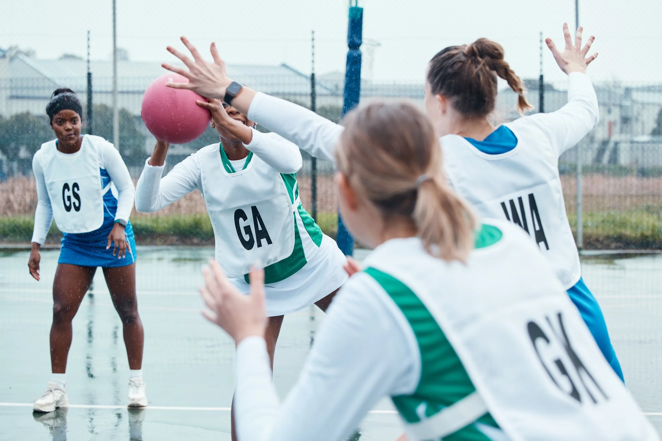 Group of women playing wheelchair basketball outside on a court, one woman with a pink ball trying to shoot, others nearby, some in motion, wearing sports jerseys with country codes, on a cloudy day.