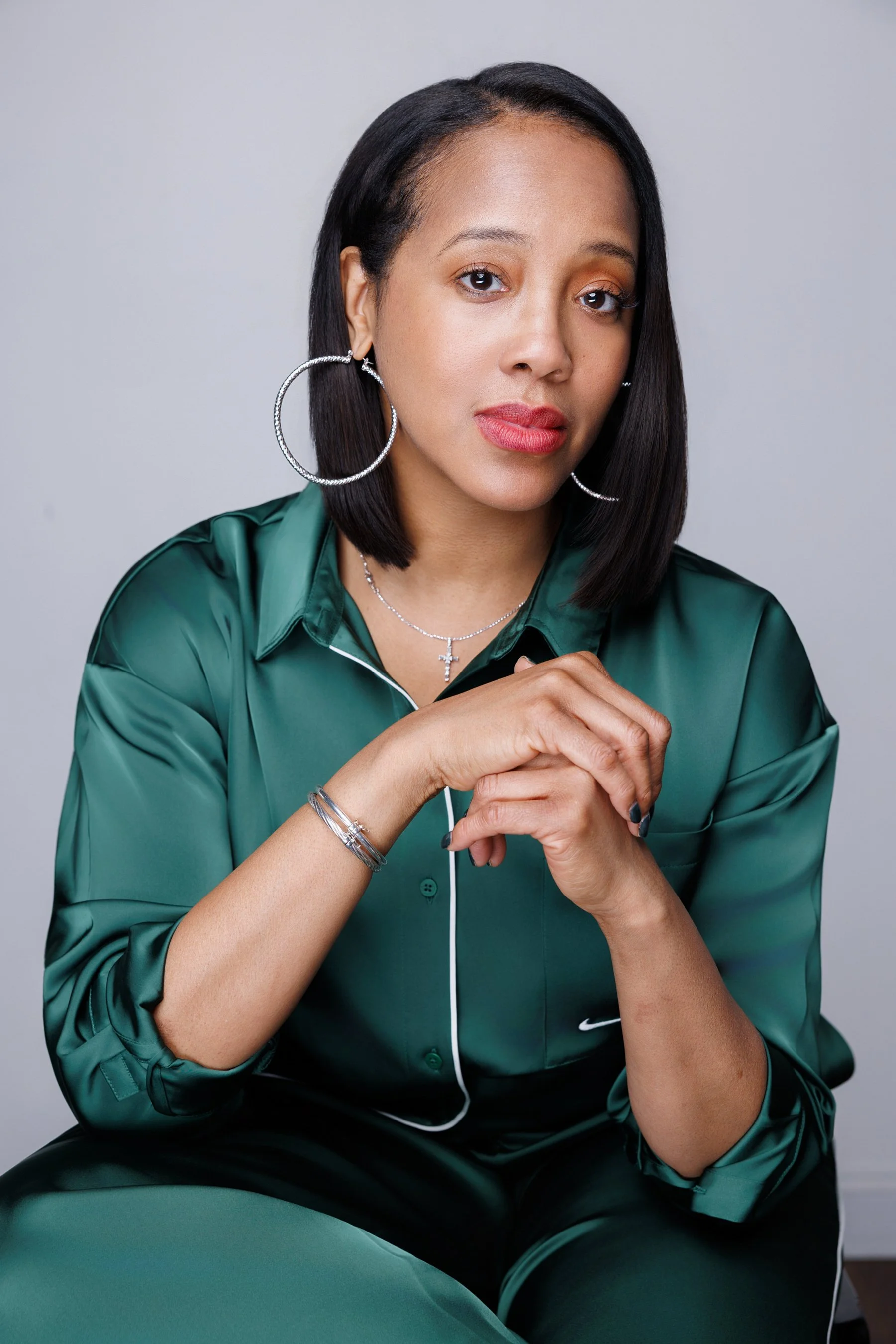 A woman with shoulder-length black hair wearing large hoop earrings, a silver cross necklace, a silver bracelet, and a dark green satin pajama-like shirt and pants sitting with her hands clasped.