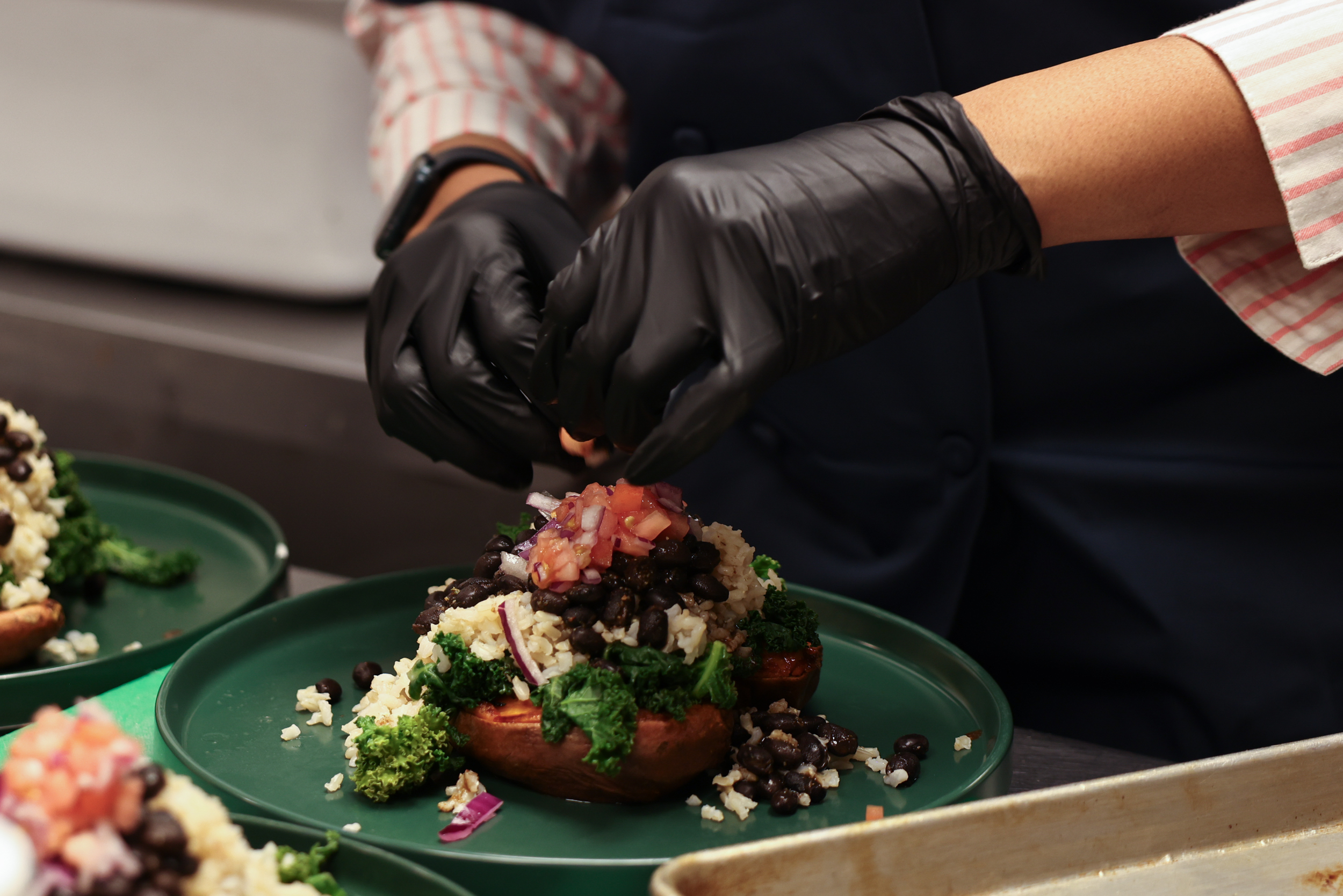 Person wearing black gloves and a striped shirt preparing a loaded baked potato with toppings such as black beans, cheese, broccoli, and salsa on a green plate.